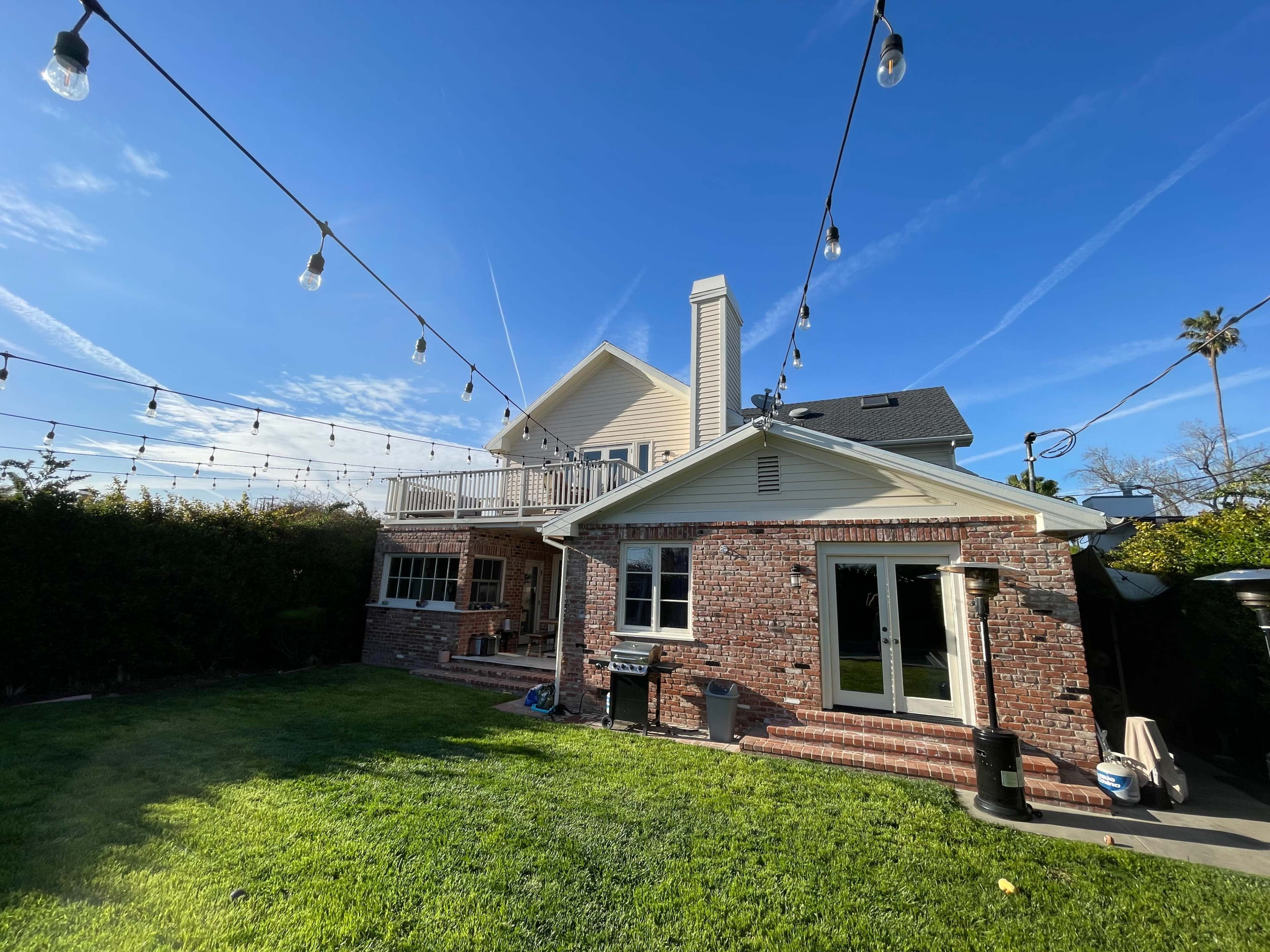 A two-story house with a brick wall and a large backyard, decorated with string lights overhead against a clear blue sky.