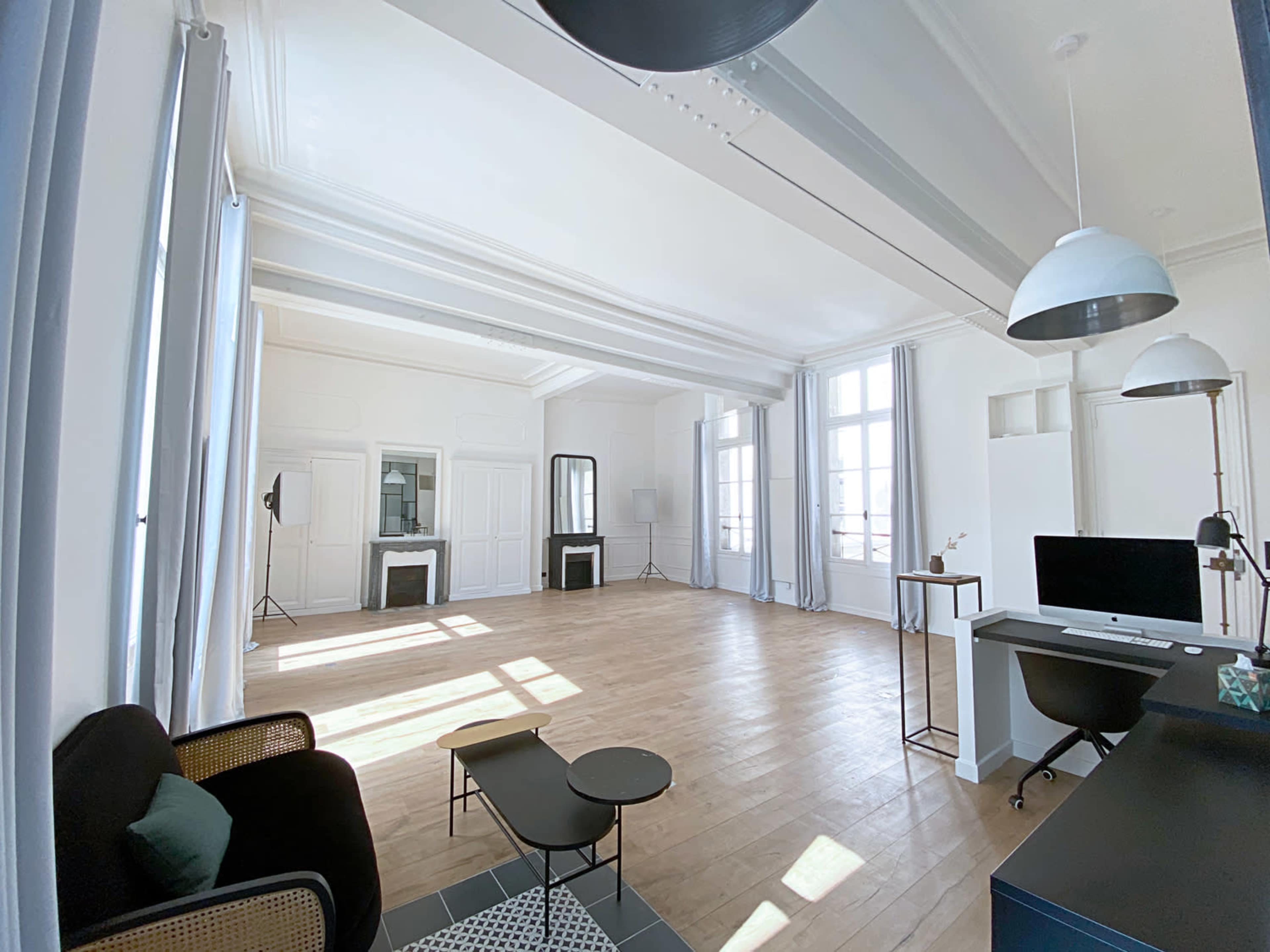 A spacious, sunlit office with light-colored walls, wooden flooring, and large windows, furnished with a black sofa, a glass coffee table, and a desk with a computer.
