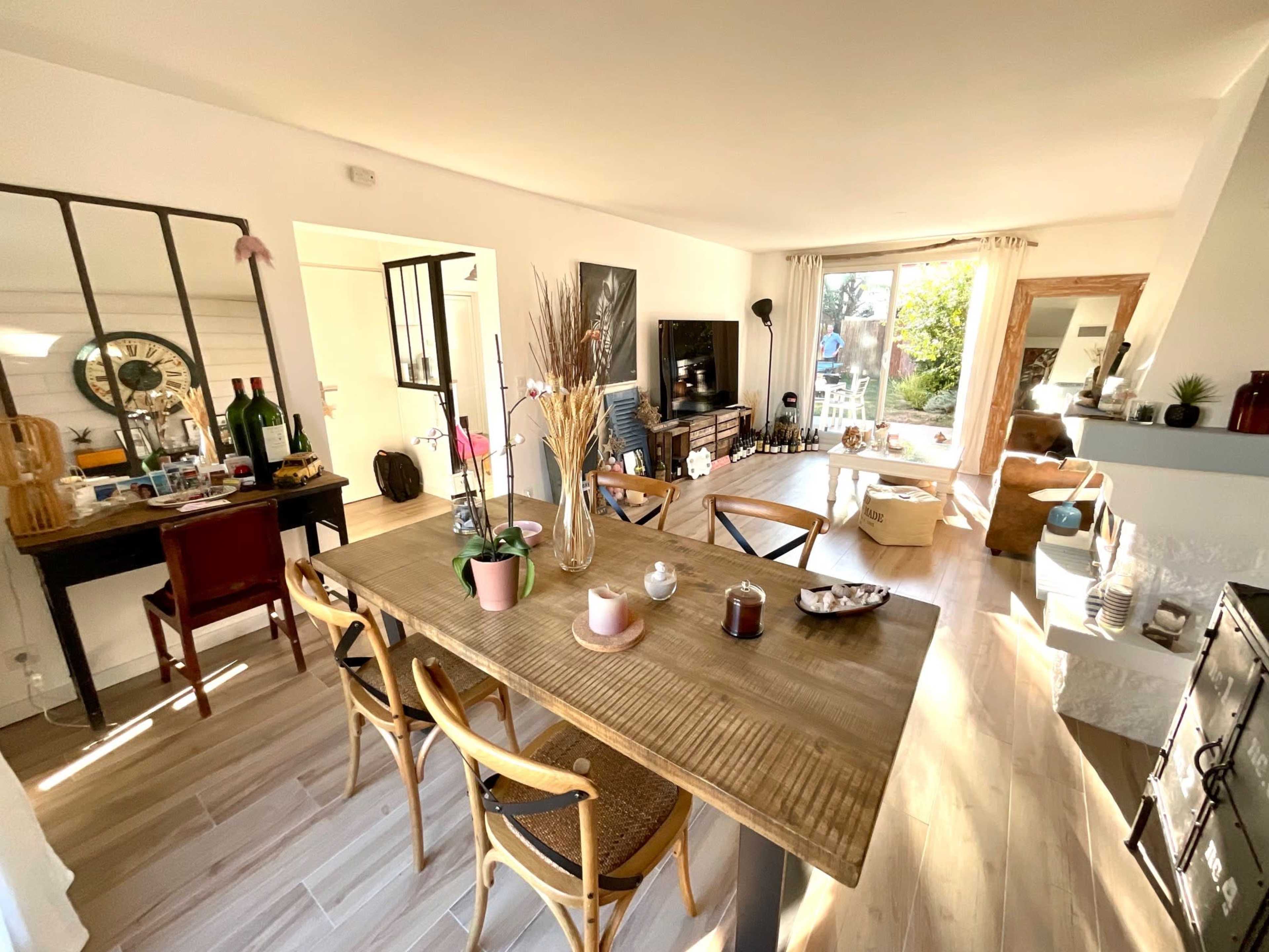 A bright living space features a wooden dining table surrounded by chairs, a desk with decor and bottles, and a cozy seating area with a fireplace and large windows.