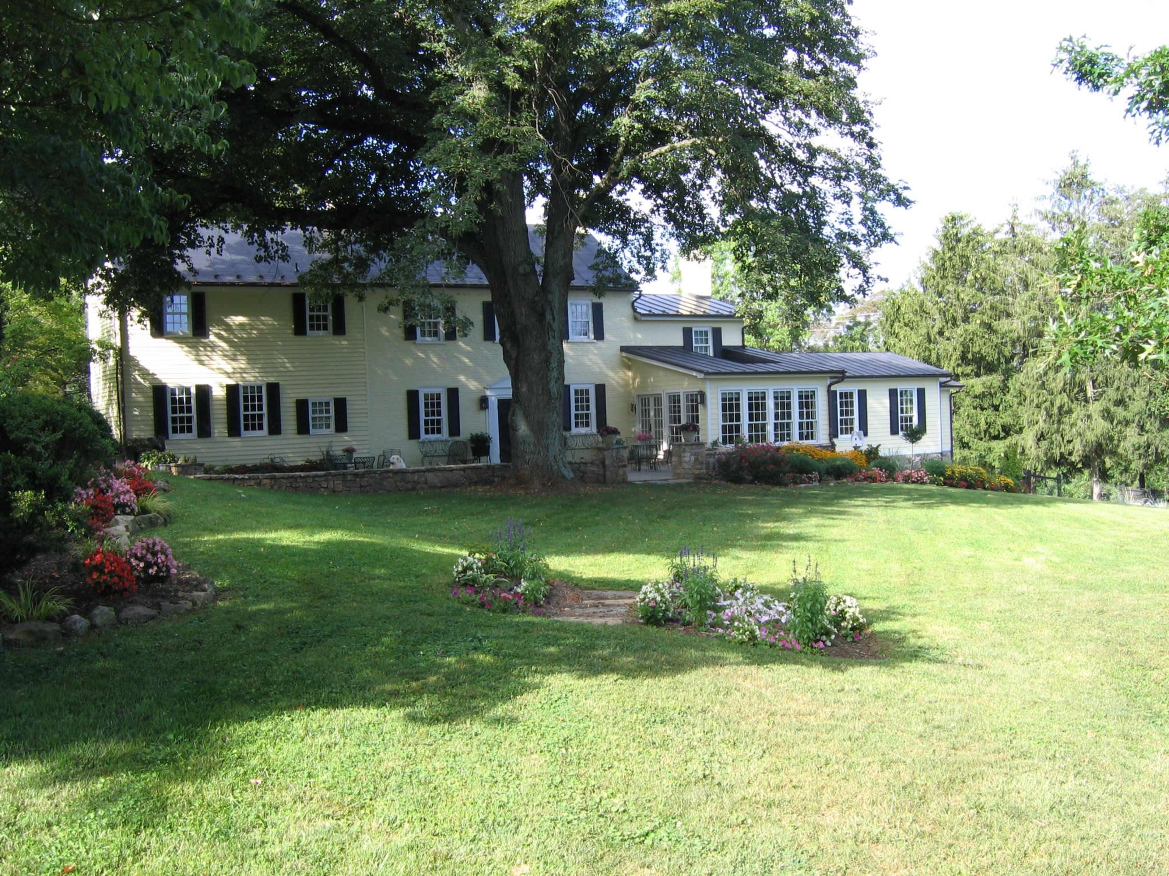 A two-story yellow house with a dark roof is surrounded by a landscaped yard featuring flower beds and large trees.