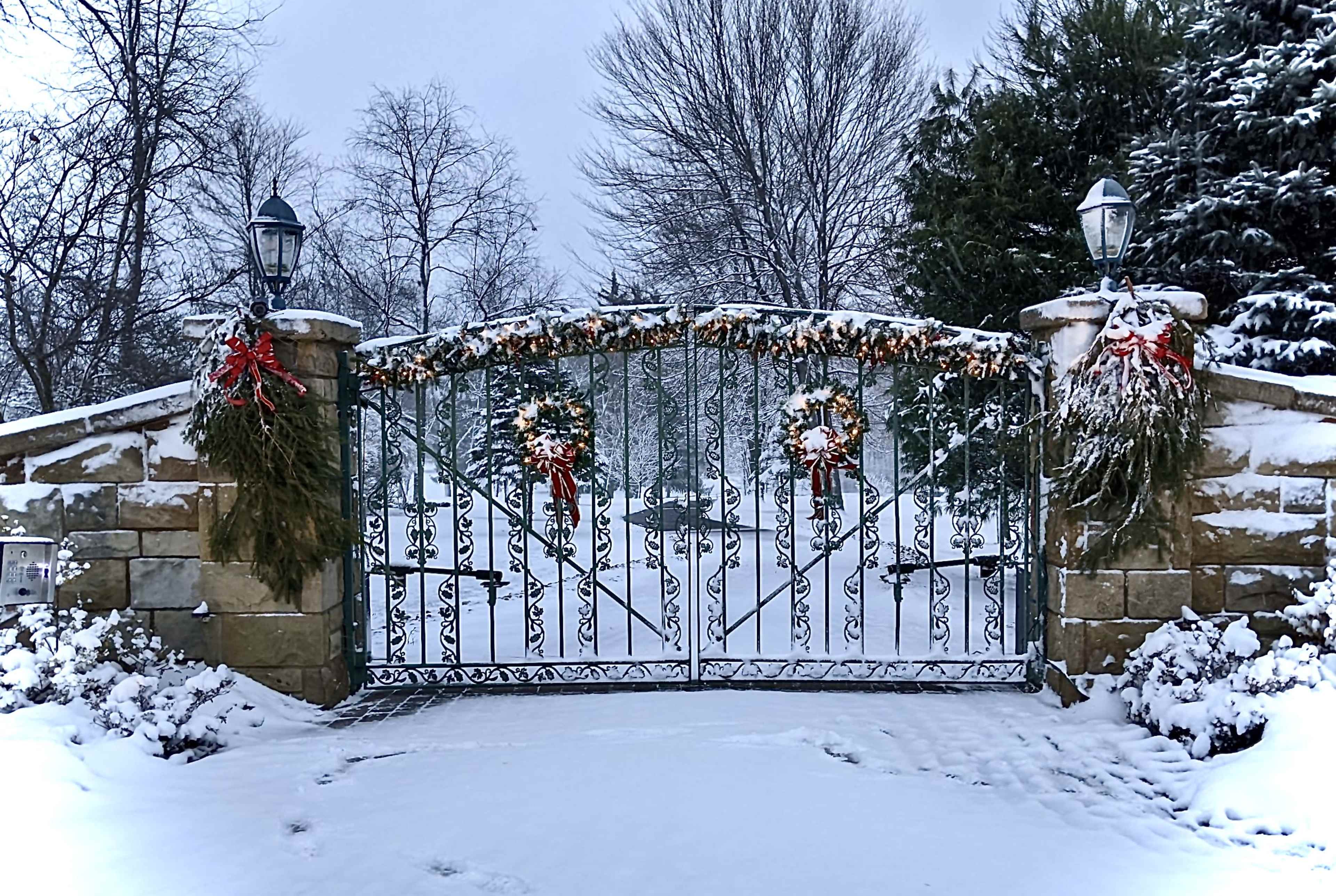 A wrought iron gate adorned with festive wreaths and garlands stands closed amidst a snowy landscape.