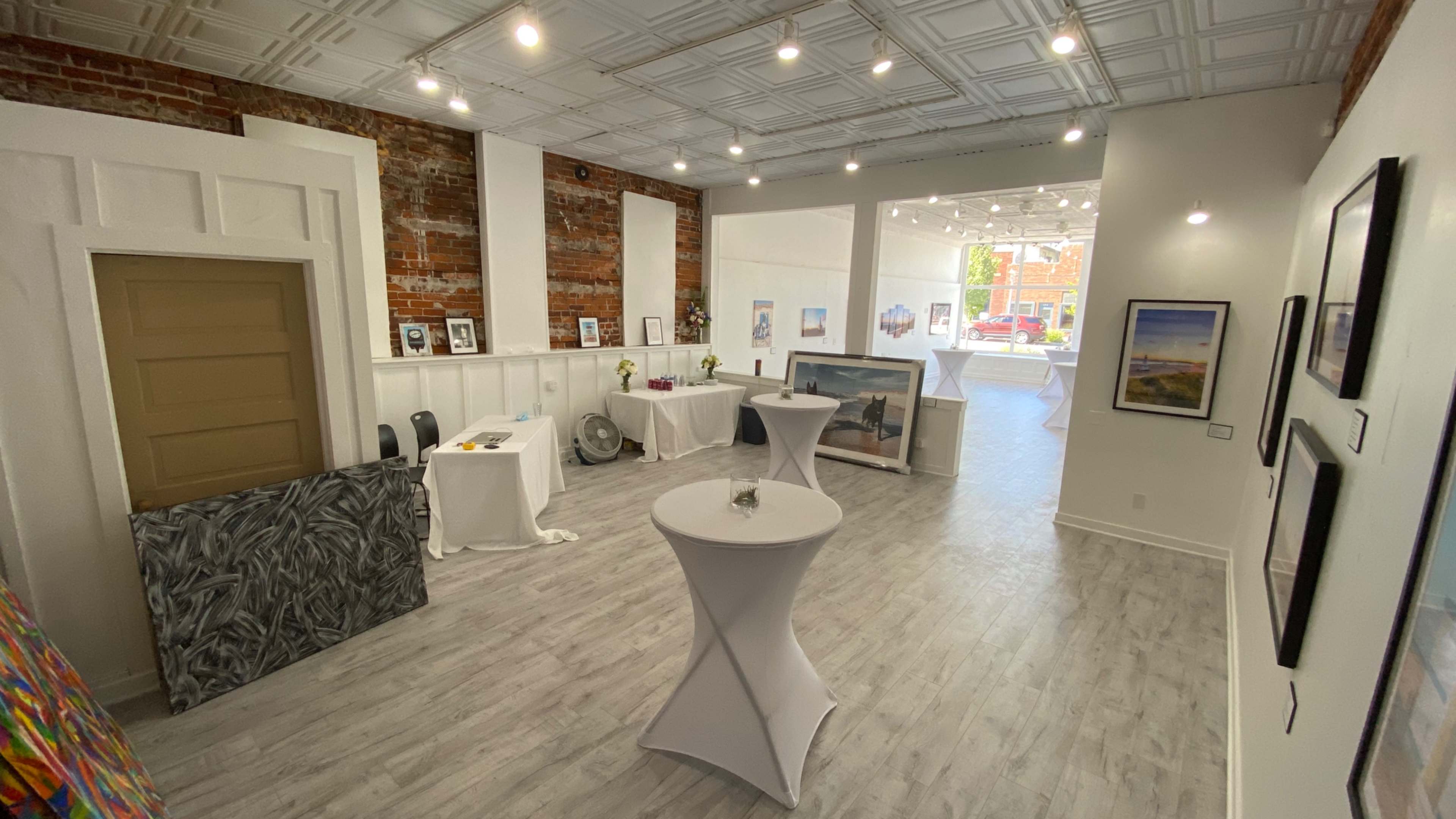 The image shows a bright art gallery with white walls, showcasing various artworks and decorative tables in an open space.