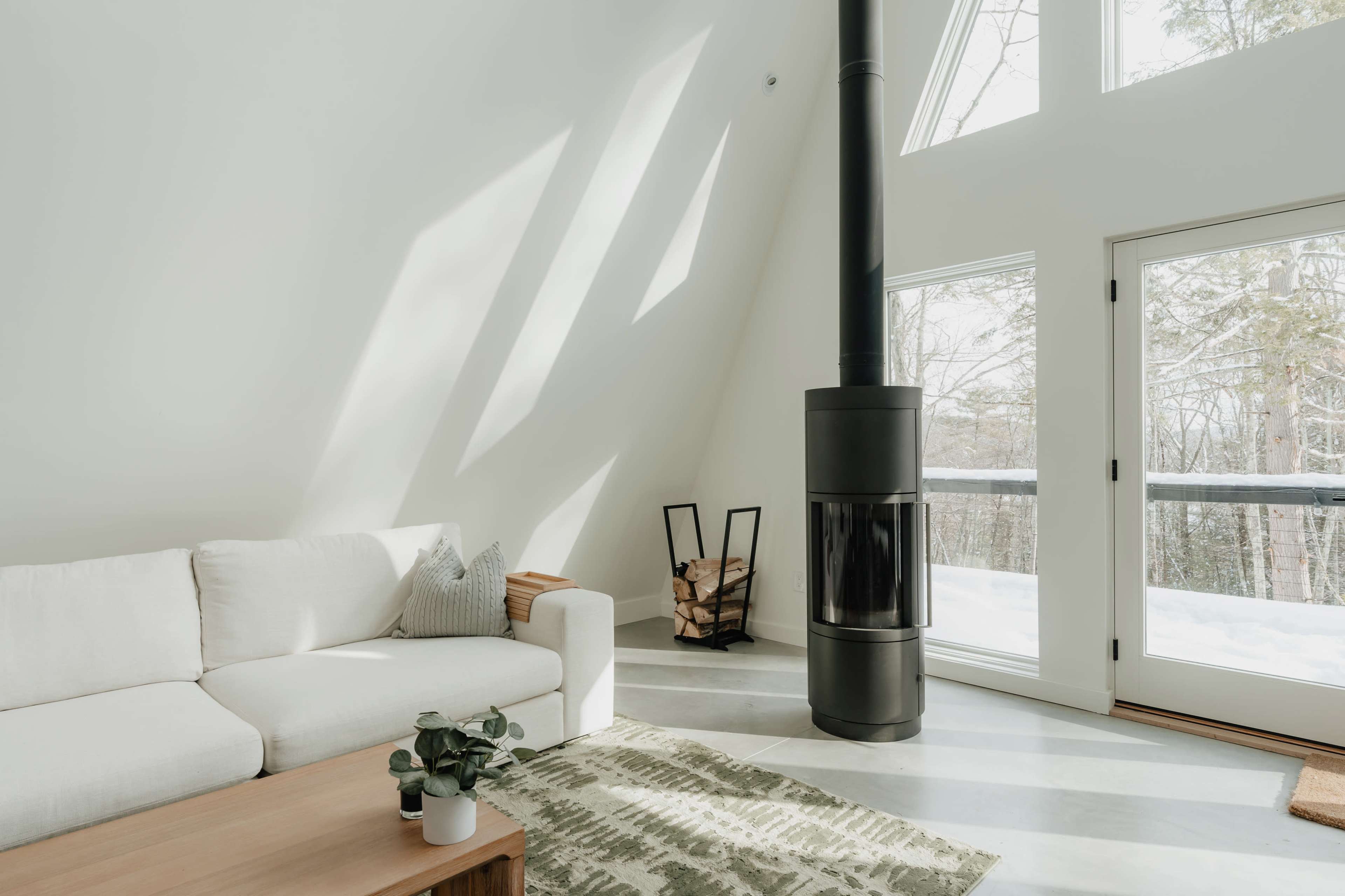 A modern living room features a sectional couch, a minimalist coffee table, a black wood stove, and large windows overlooking a snowy landscape.
