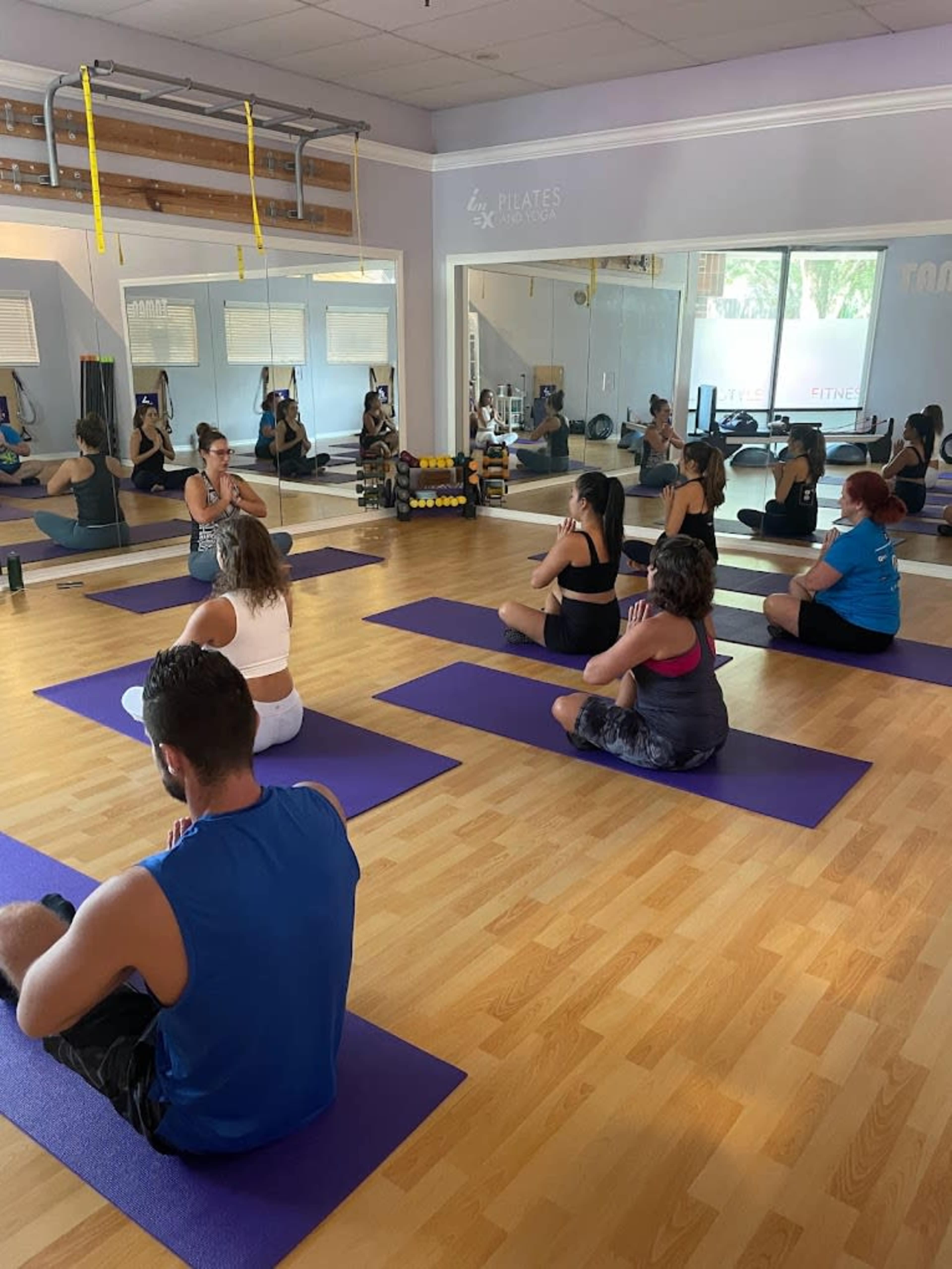 A group of people is practicing yoga on purple mats in a well-lit studio with mirrors and fitness equipment in the background.