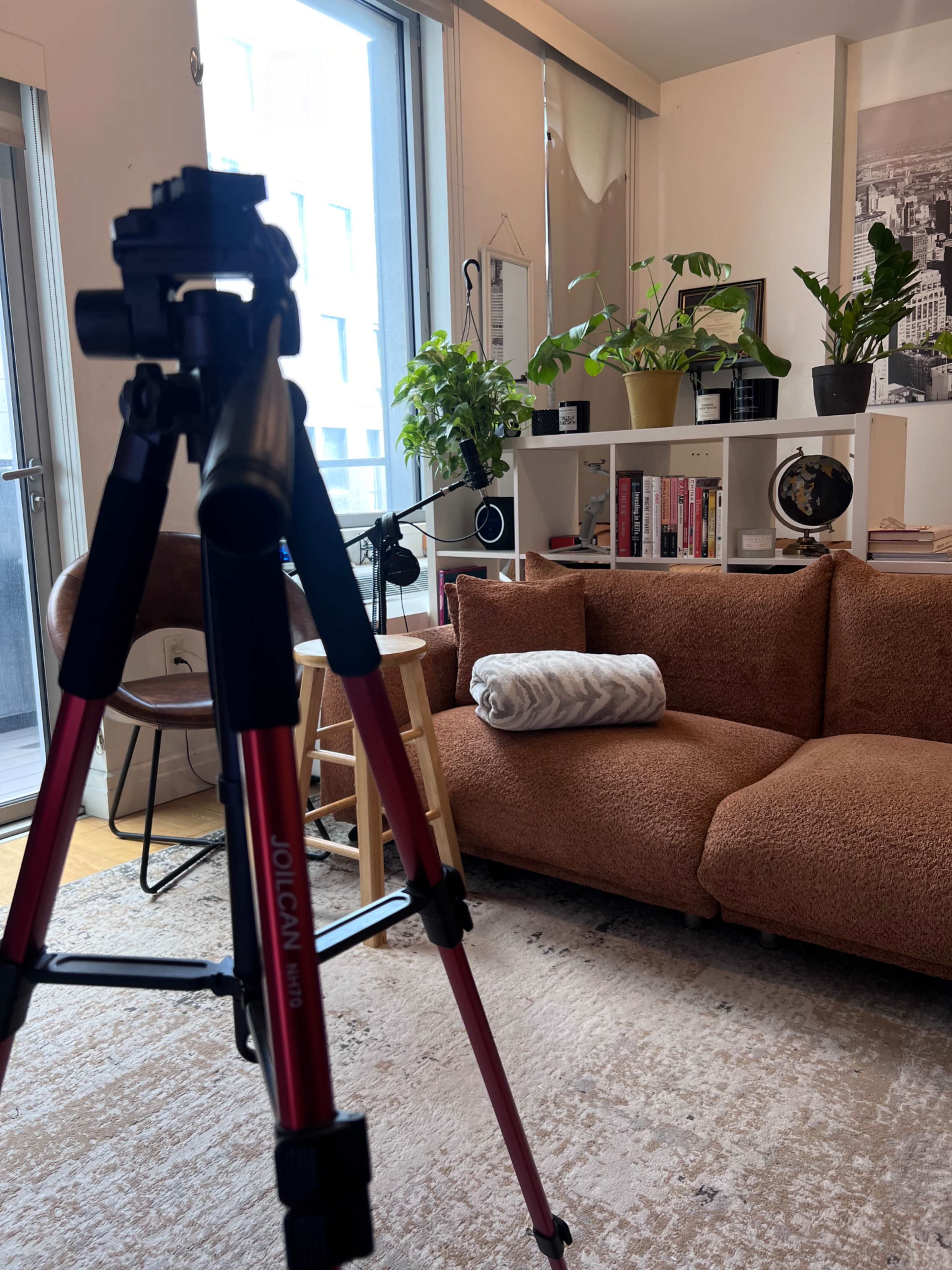 A living room with a tripod in the foreground, a cozy brown sofa, a small stool, and plants on shelves.