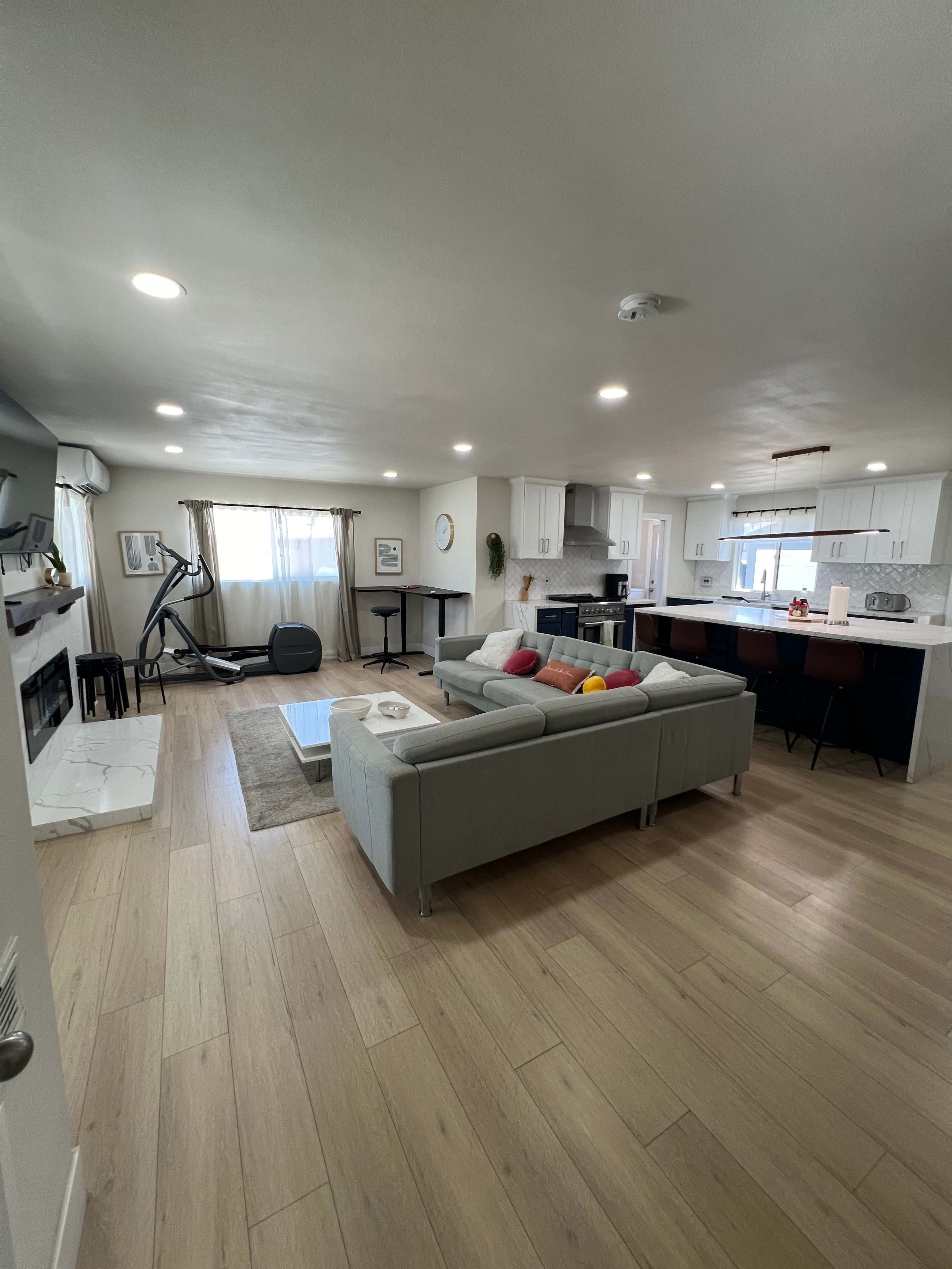 A modern living room with a sofa, a coffee table, a fitness machine, and a kitchen area in the background.