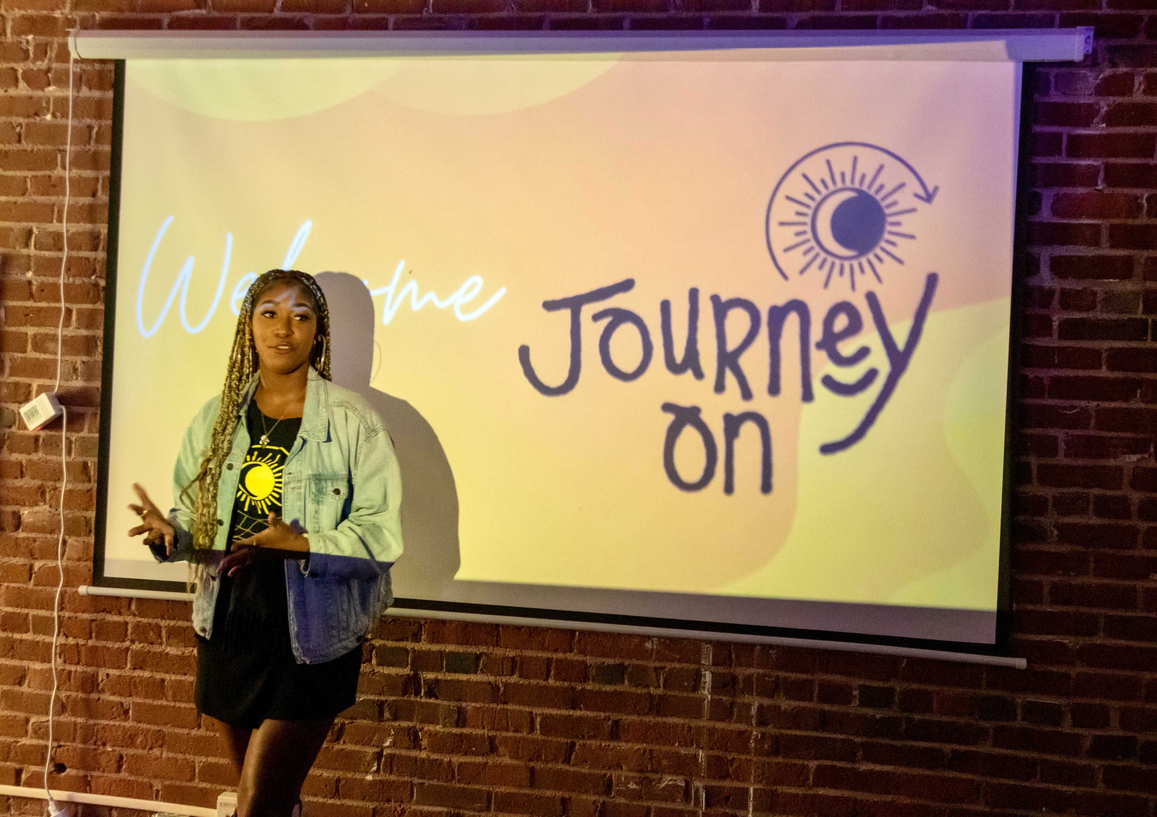 A woman stands in front of a projector screen displaying the words "Welcome" and "Journey On" against a vibrant background in a brick-walled room.