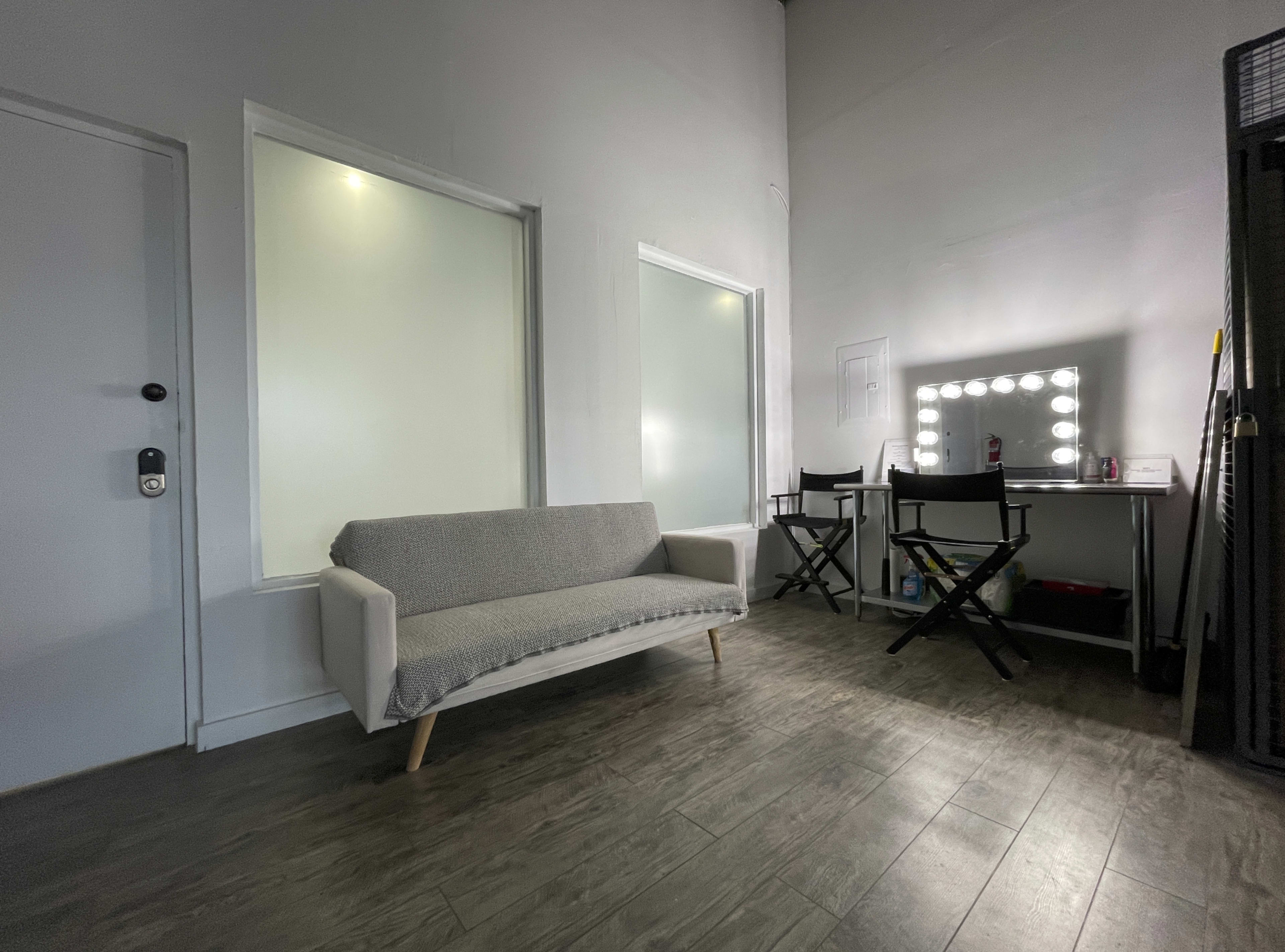 A small room with a gray sofa and a makeup station featuring a mirror with bright lights, along with two black folding chairs.