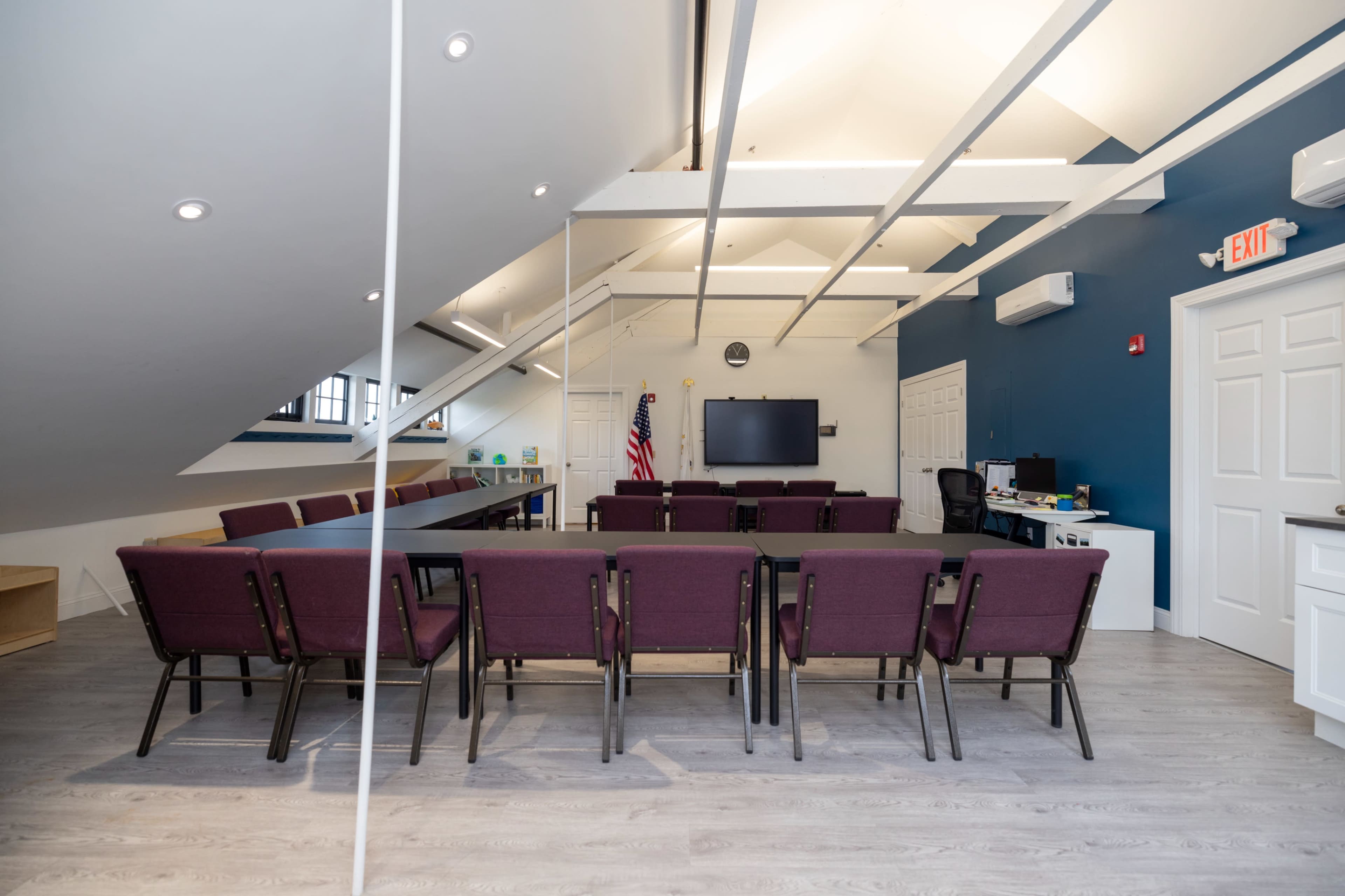 A conference room with a long table surrounded by maroon chairs, a television on the wall, and a large window in an attic space with sloped ceilings.