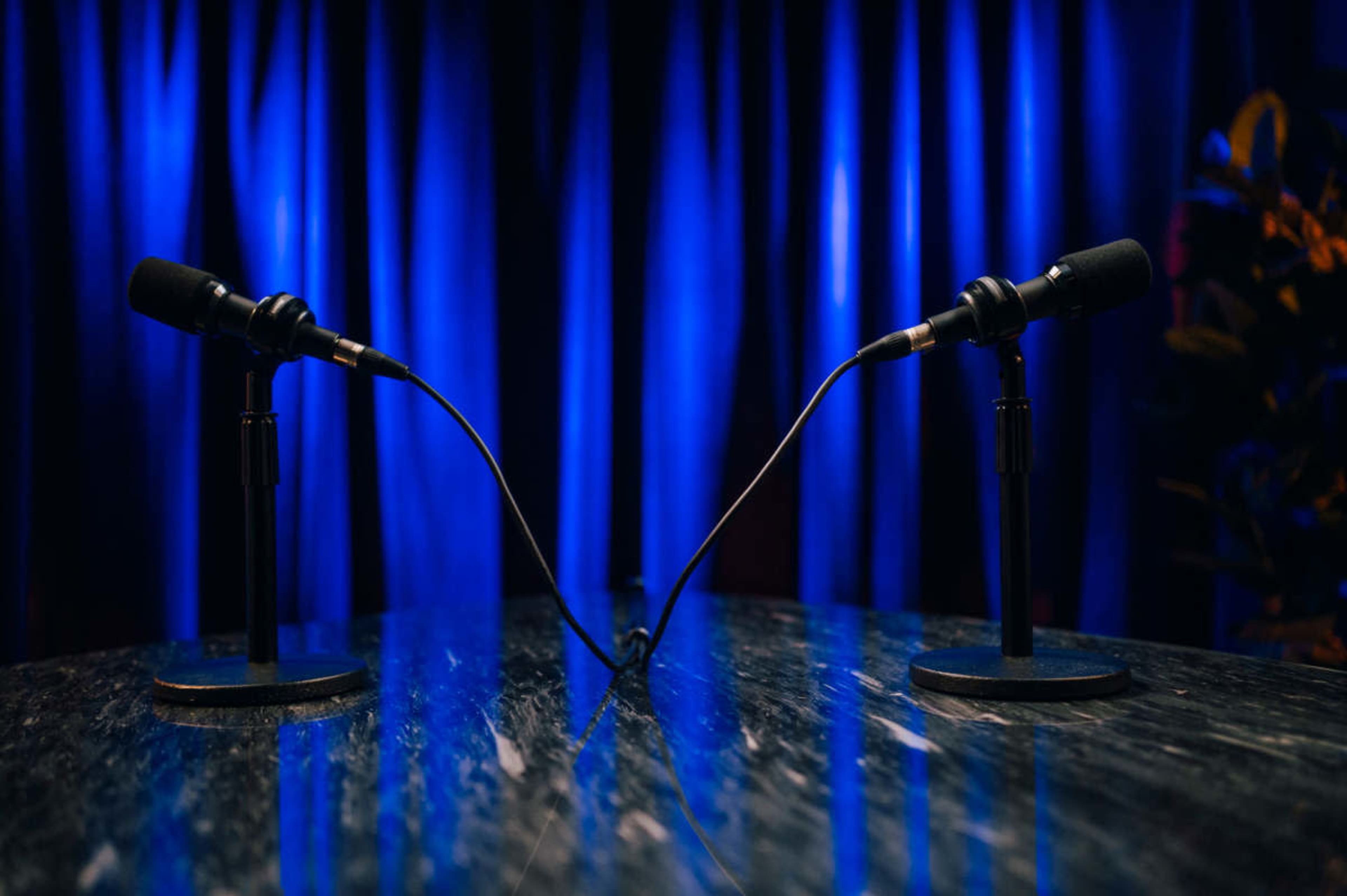 Two microphones stand on a tabletop facing each other, with a blue curtain draped in the background.