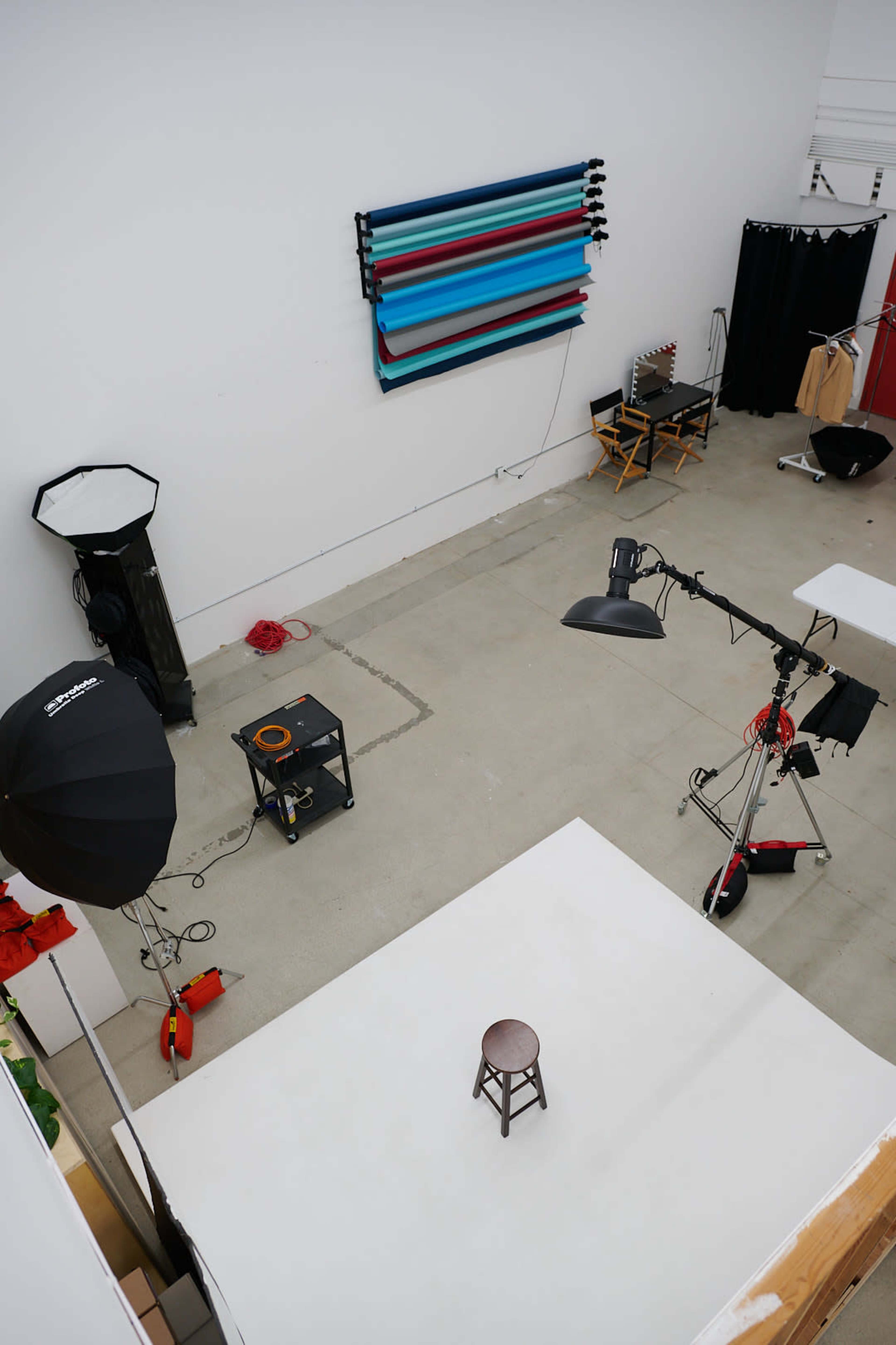 The image shows an empty photography studio with a white backdrop, a stool in the center, and various lighting equipment arranged around the space.