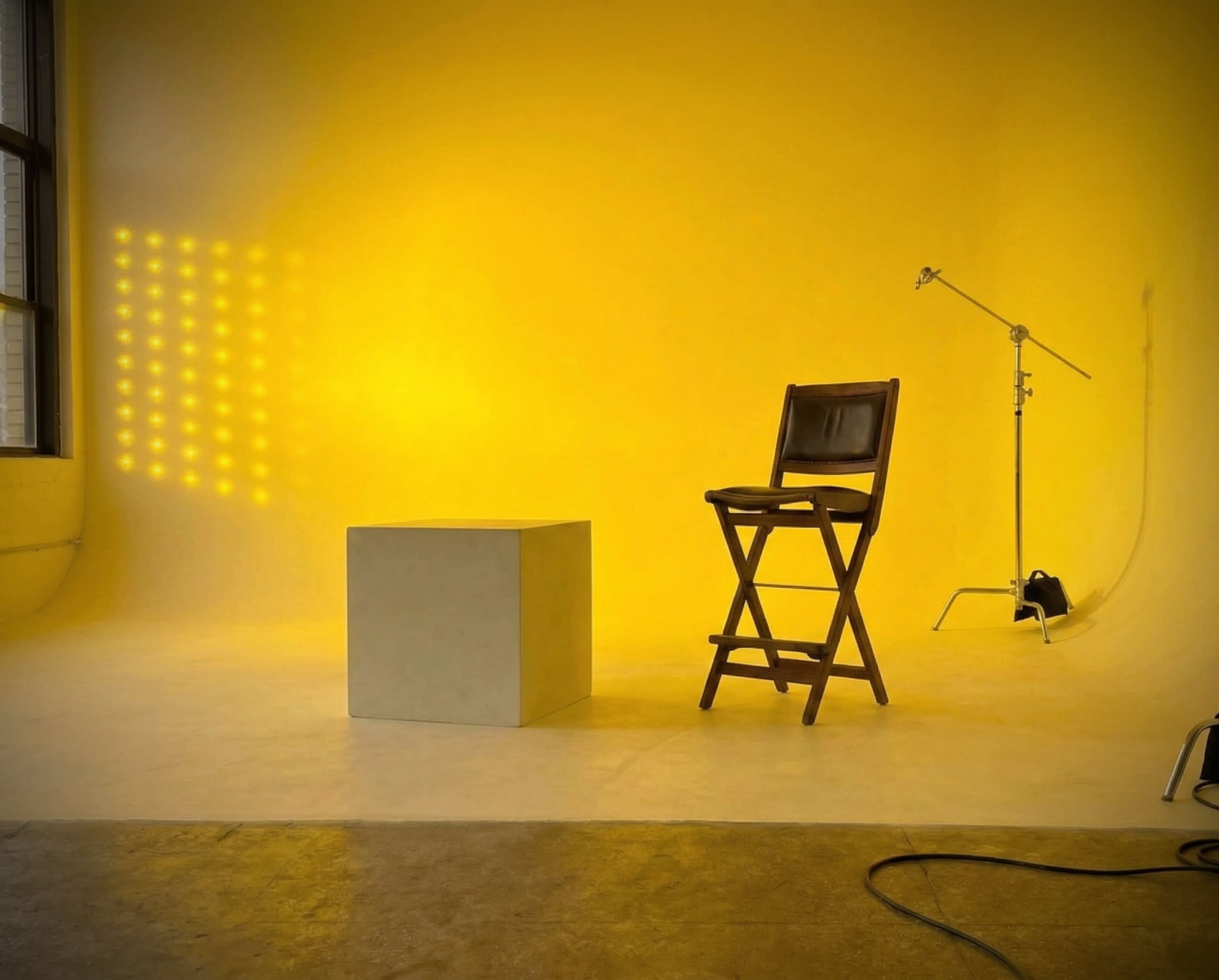 The image shows a minimalistic studio setup with a wooden chair and a cubic pedestal against a vibrant yellow backdrop.
