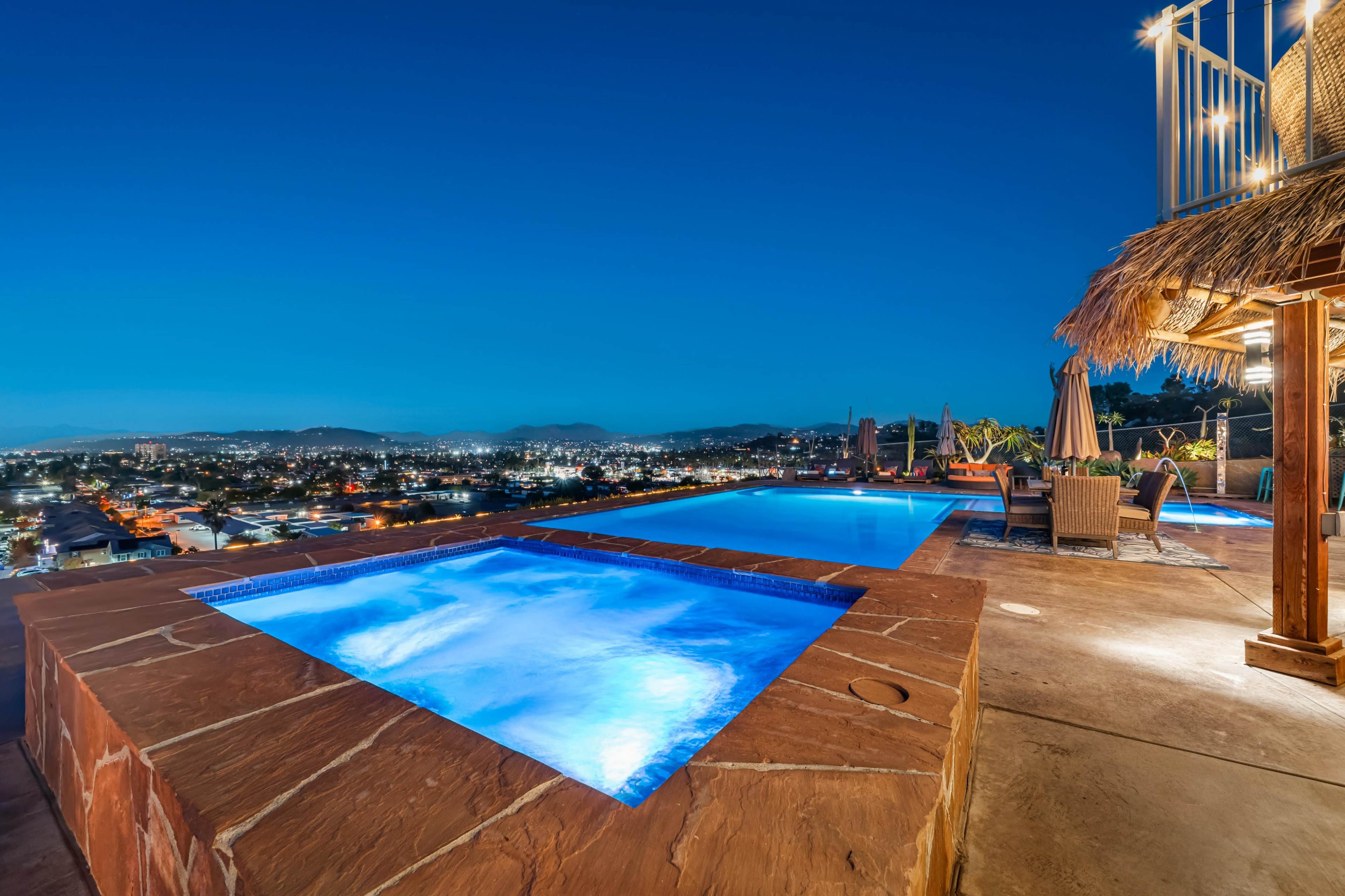 A rooftop area features two illuminated swimming pools with a view of a city skyline at twilight.