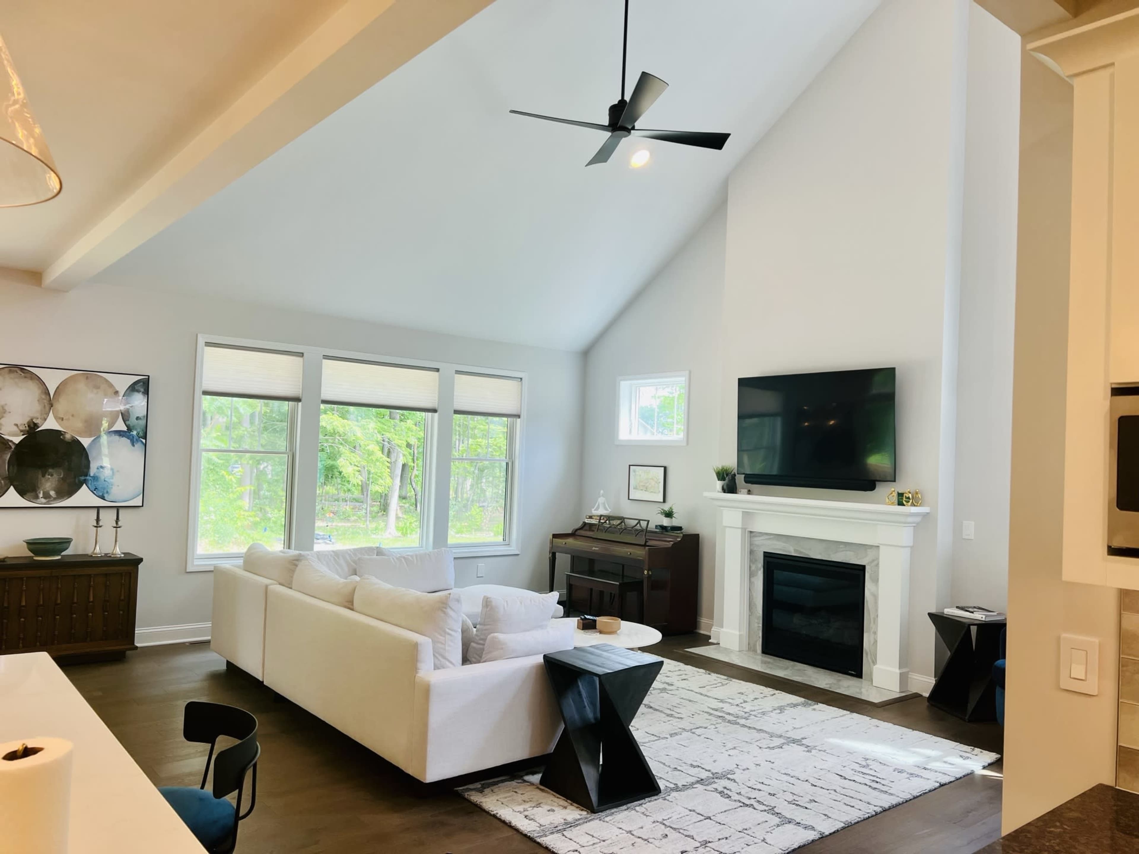 A modern living room features a white sectional sofa facing a large television above a fireplace, with large windows allowing natural light to illuminate the space.
