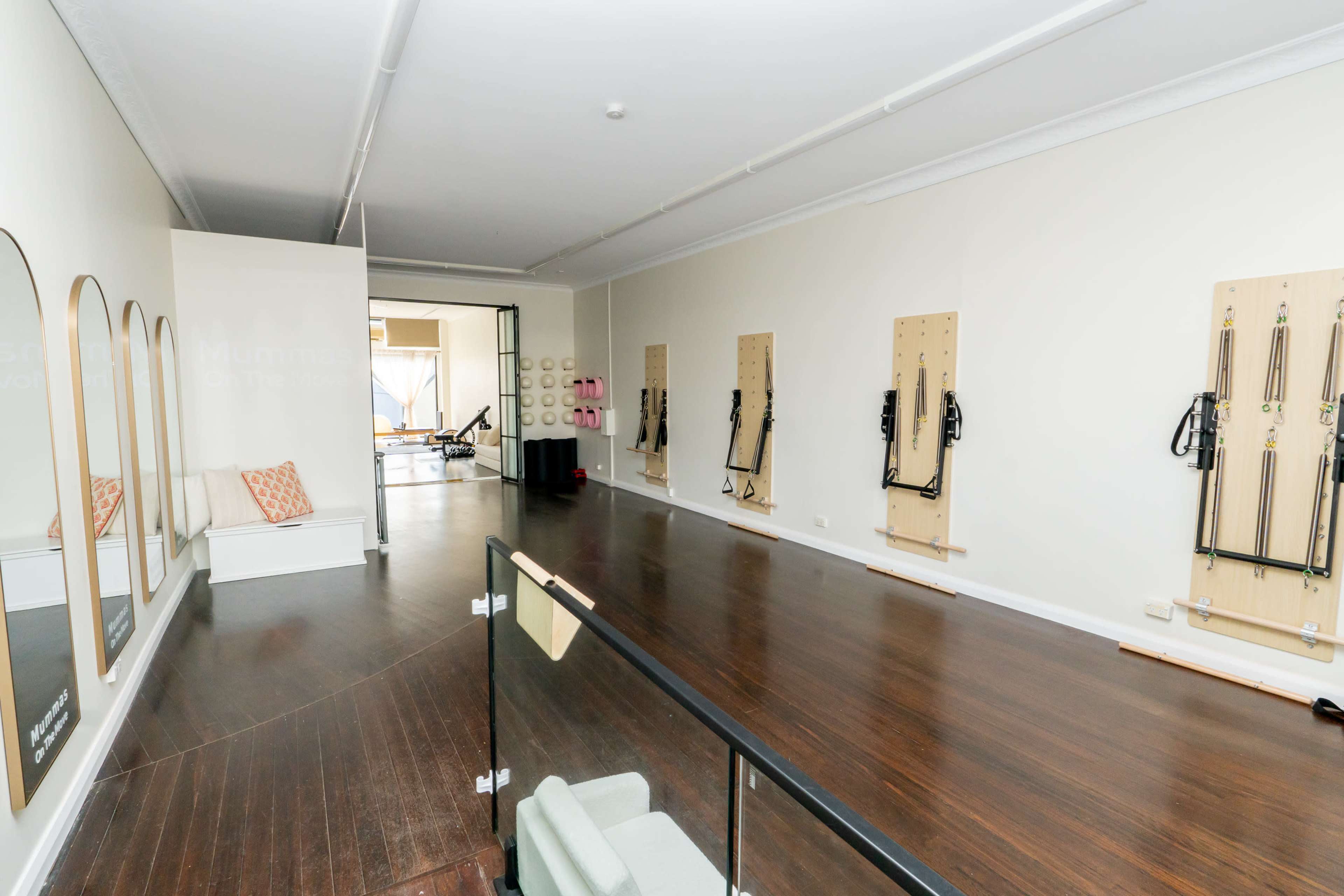 The image shows a spacious, well-lit studio with wooden flooring, featuring multiple wall-mounted exercise equipment stations and minimal furnishings.