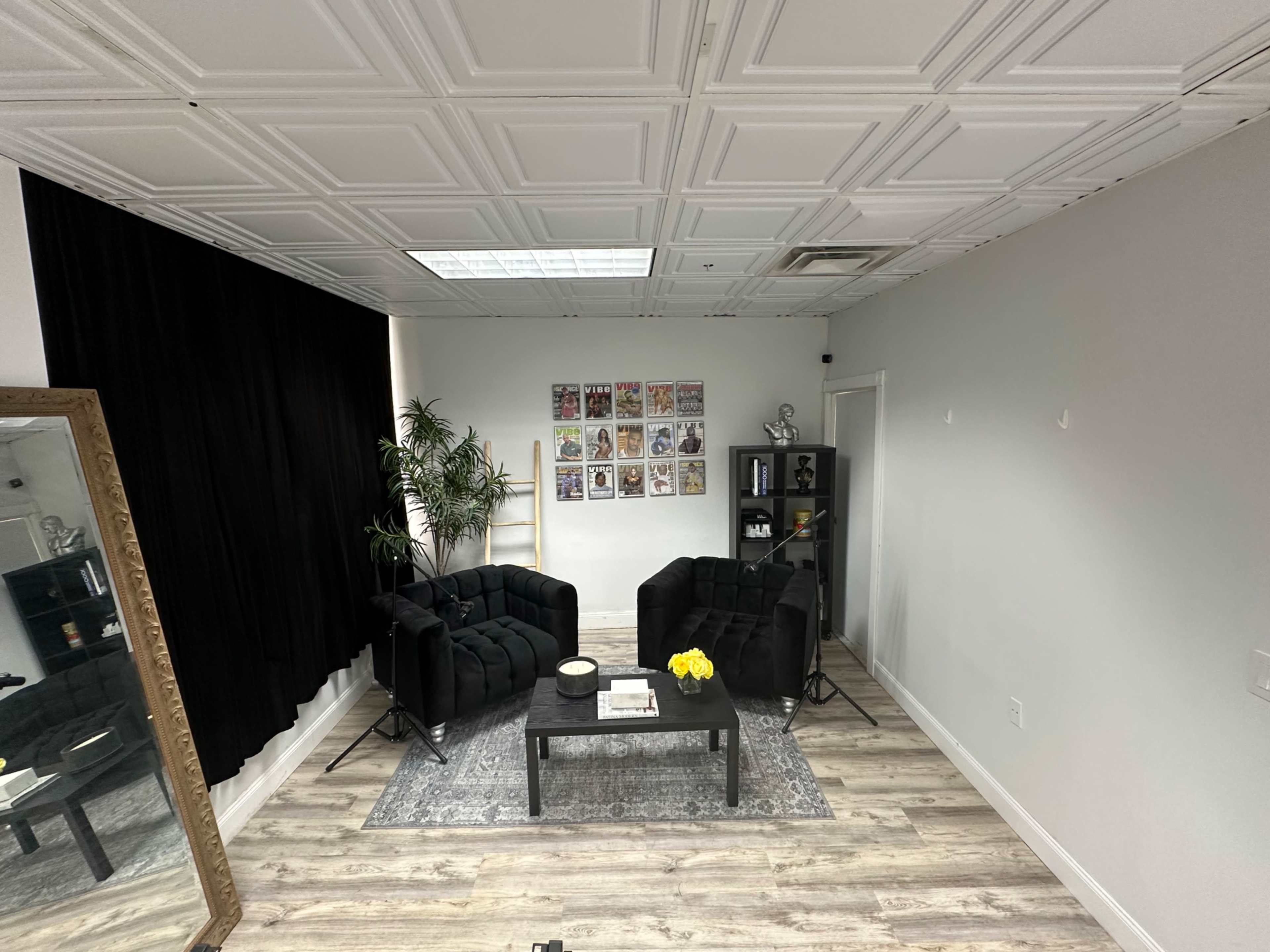 The image shows a modern lounge area with two black sofas facing a coffee table, surrounded by decor including a plant and a wall-hung collection of framed pictures.