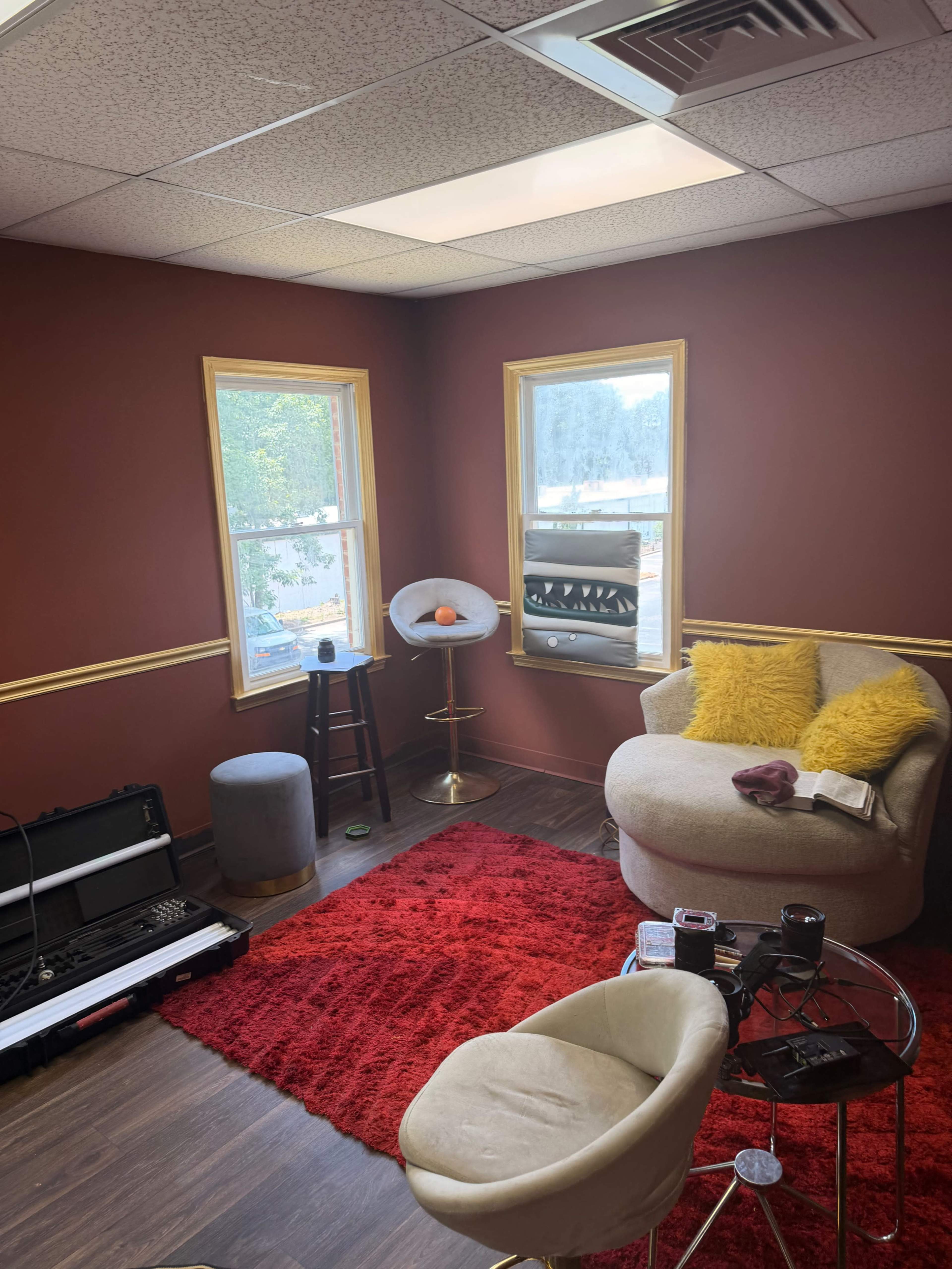 The image shows a cozy room with a plush chair, a small table, and large windows, featuring a red rug and beige walls with yellow accents.