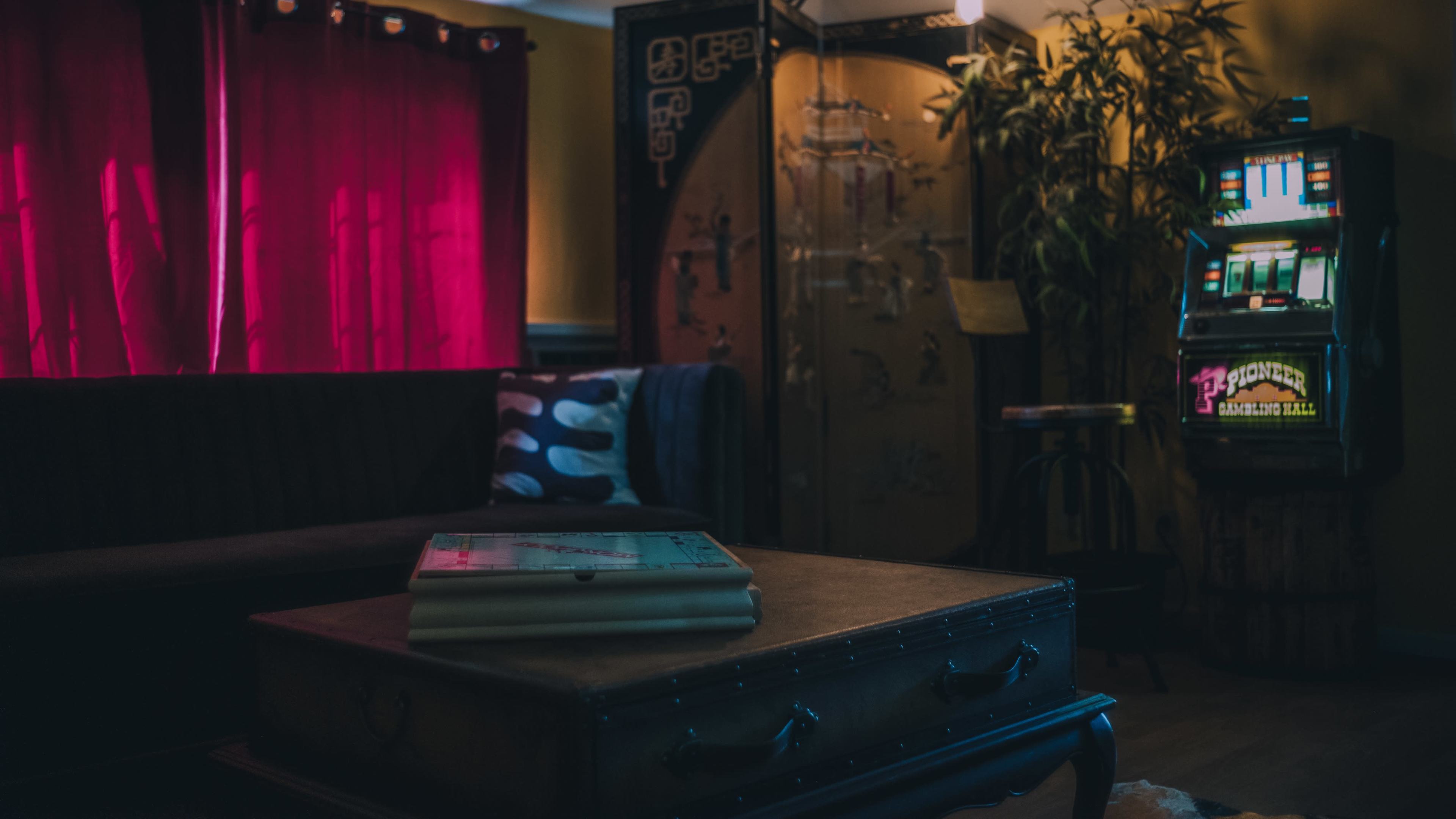 The room features a dark-colored sofa, a coffee table with books, red curtains, a decorative wall, and a vintage slot machine.