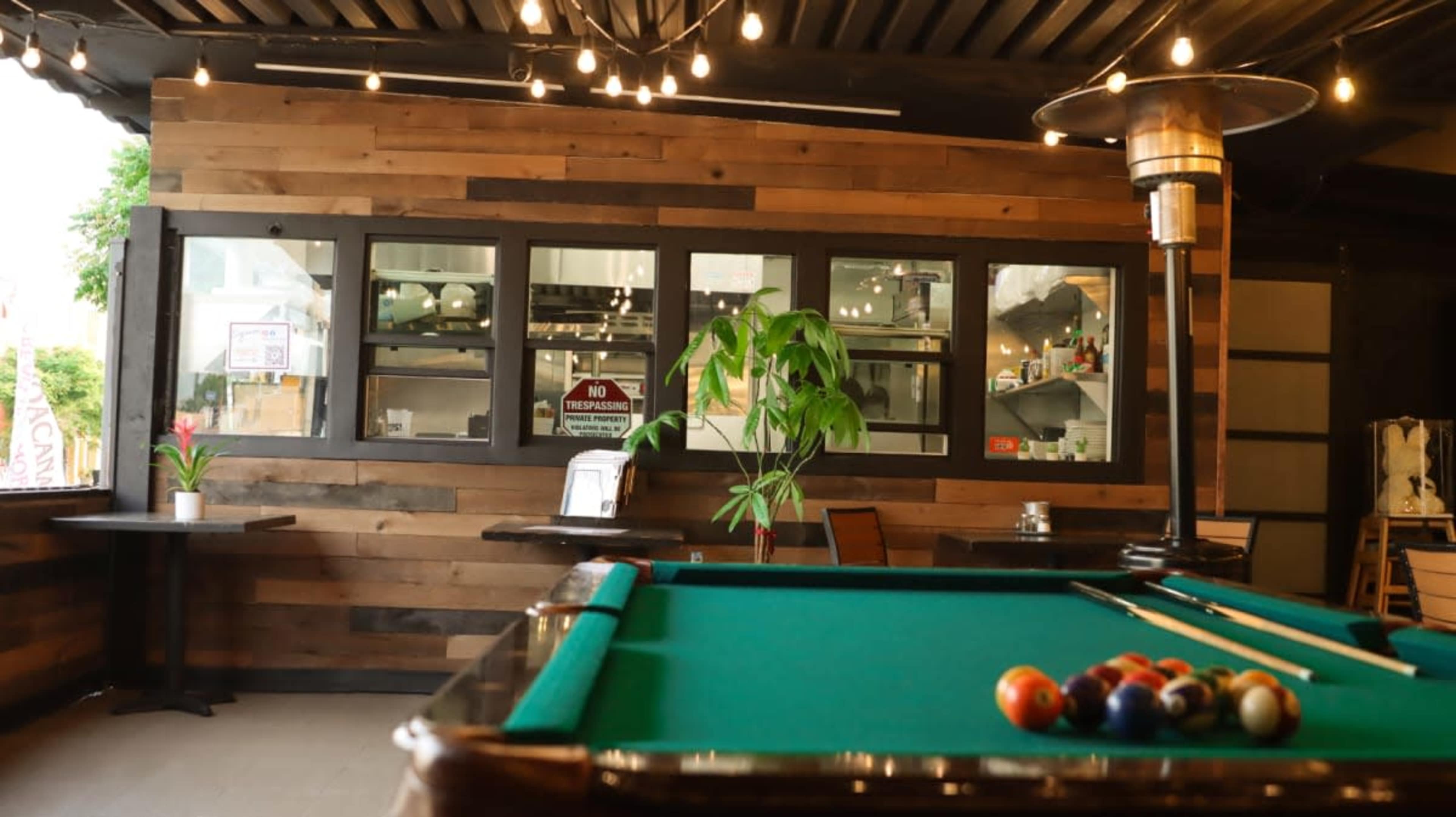 The image shows a billiard table with colorful balls arranged on it, located in a rustic room with wooden walls and warm lighting.