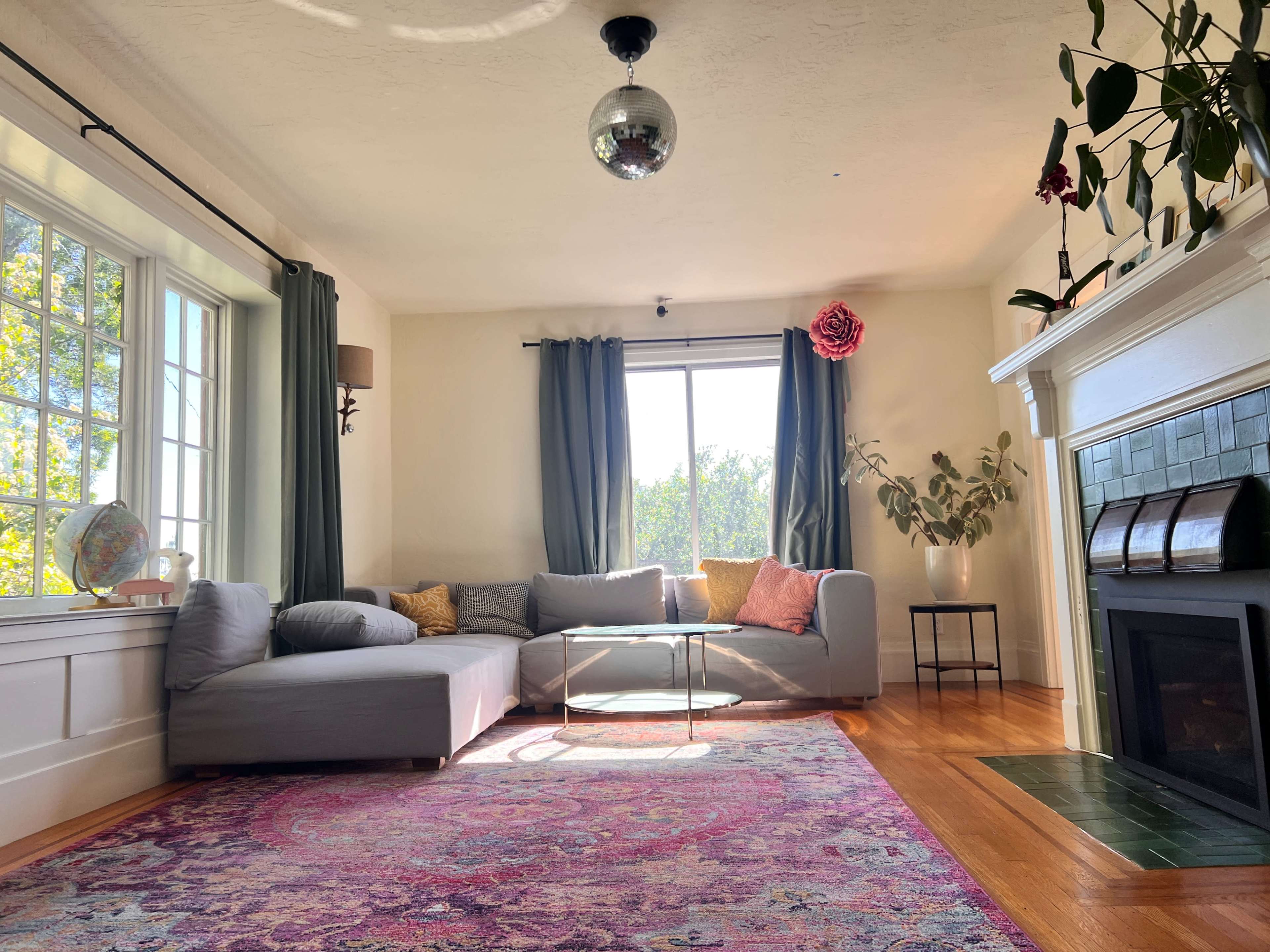 A spacious living room features a gray sectional sofa, a glass coffee table, a fireplace, and large windows with green curtains, illuminated by natural light.