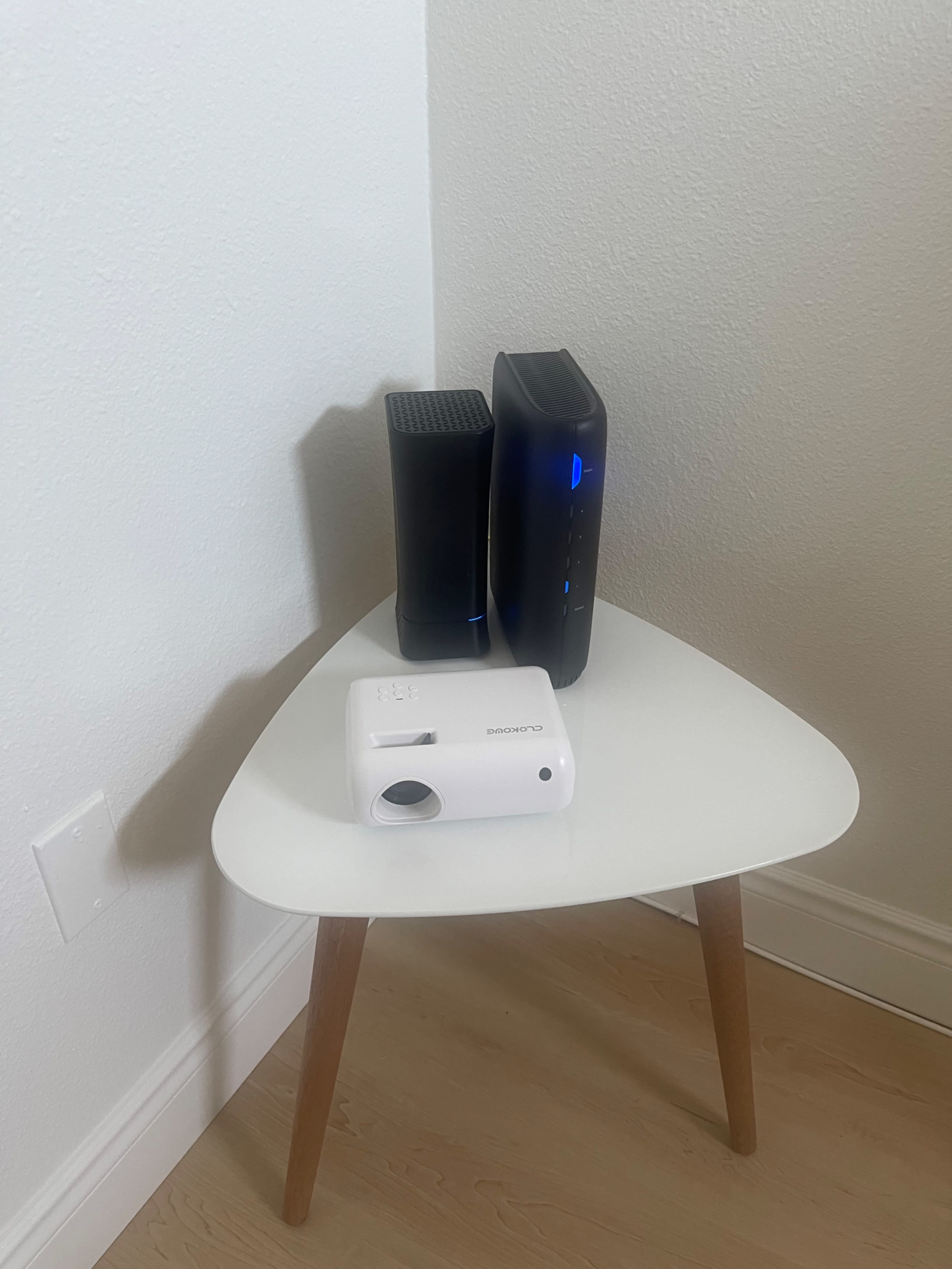 A white projector is positioned on a small, white table alongside a black modem and a black device with a blue light.