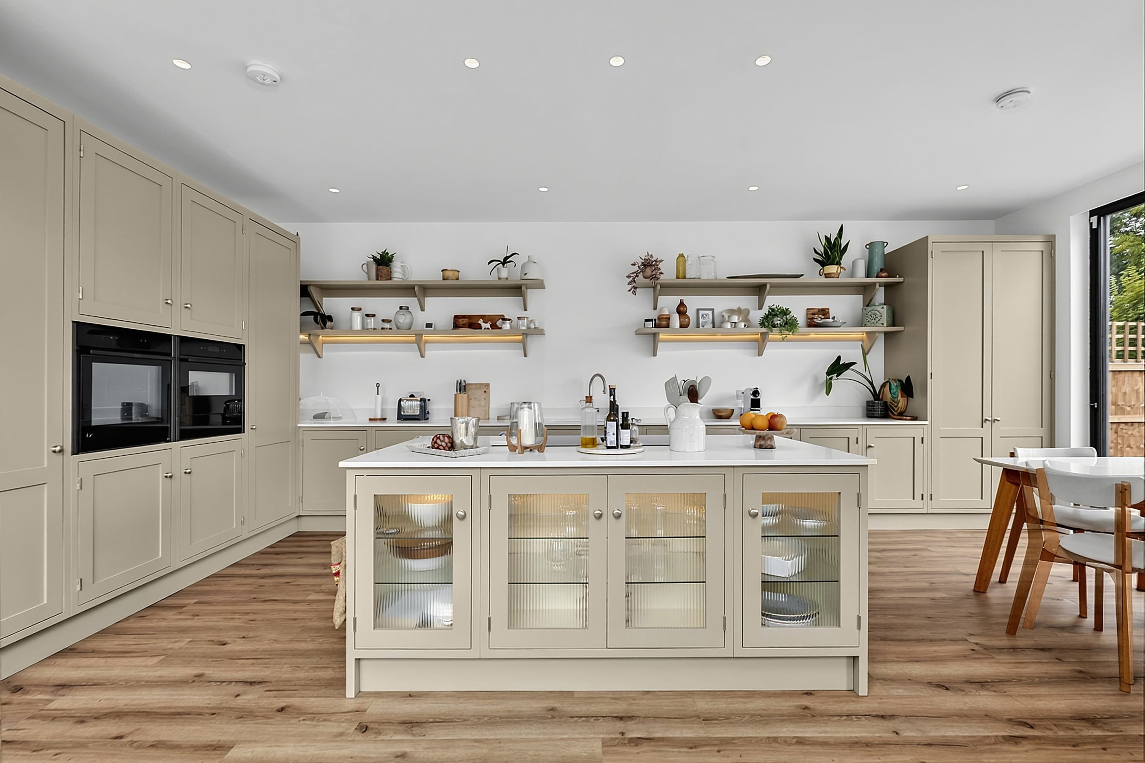 The image shows a modern kitchen featuring light-colored cabinetry, a central island with glass-front cabinets, and wooden shelves displaying various decorative items.