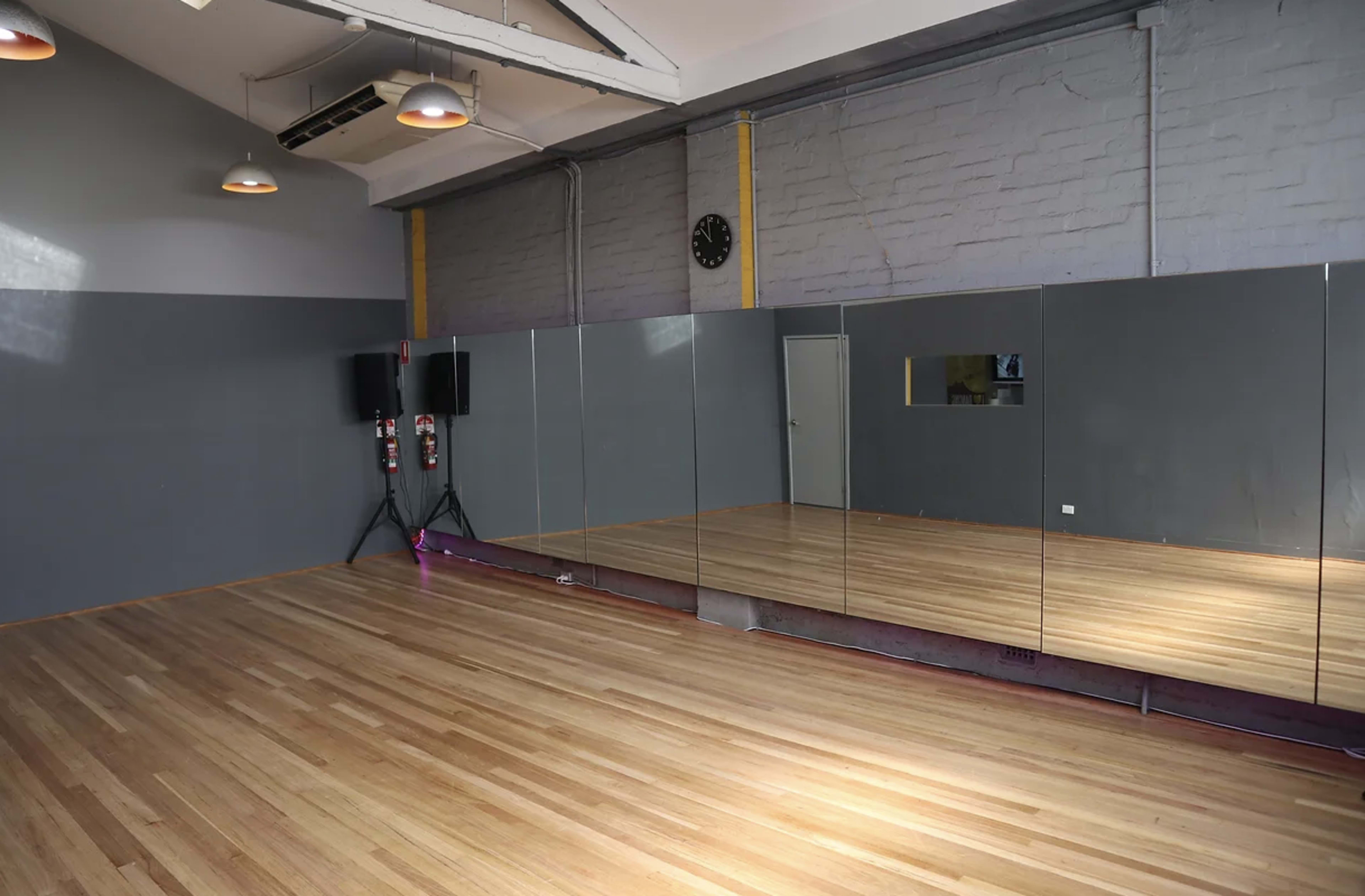 A spacious dance studio with wooden flooring, large mirrors on one wall, and minimalistic decor.