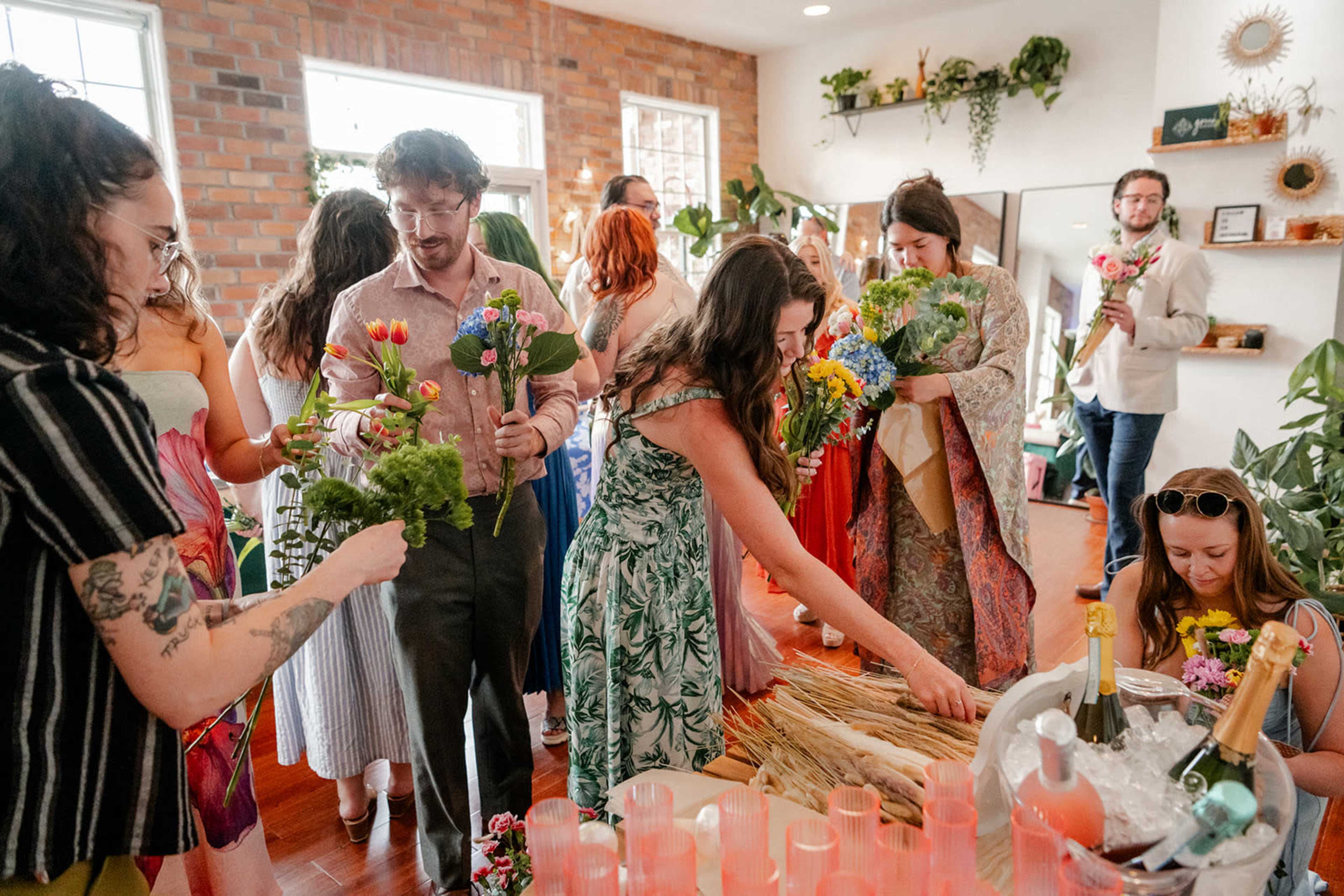A group of people gathers in a brightly lit room filled with plants, arranging flowers and drinks for a celebration.