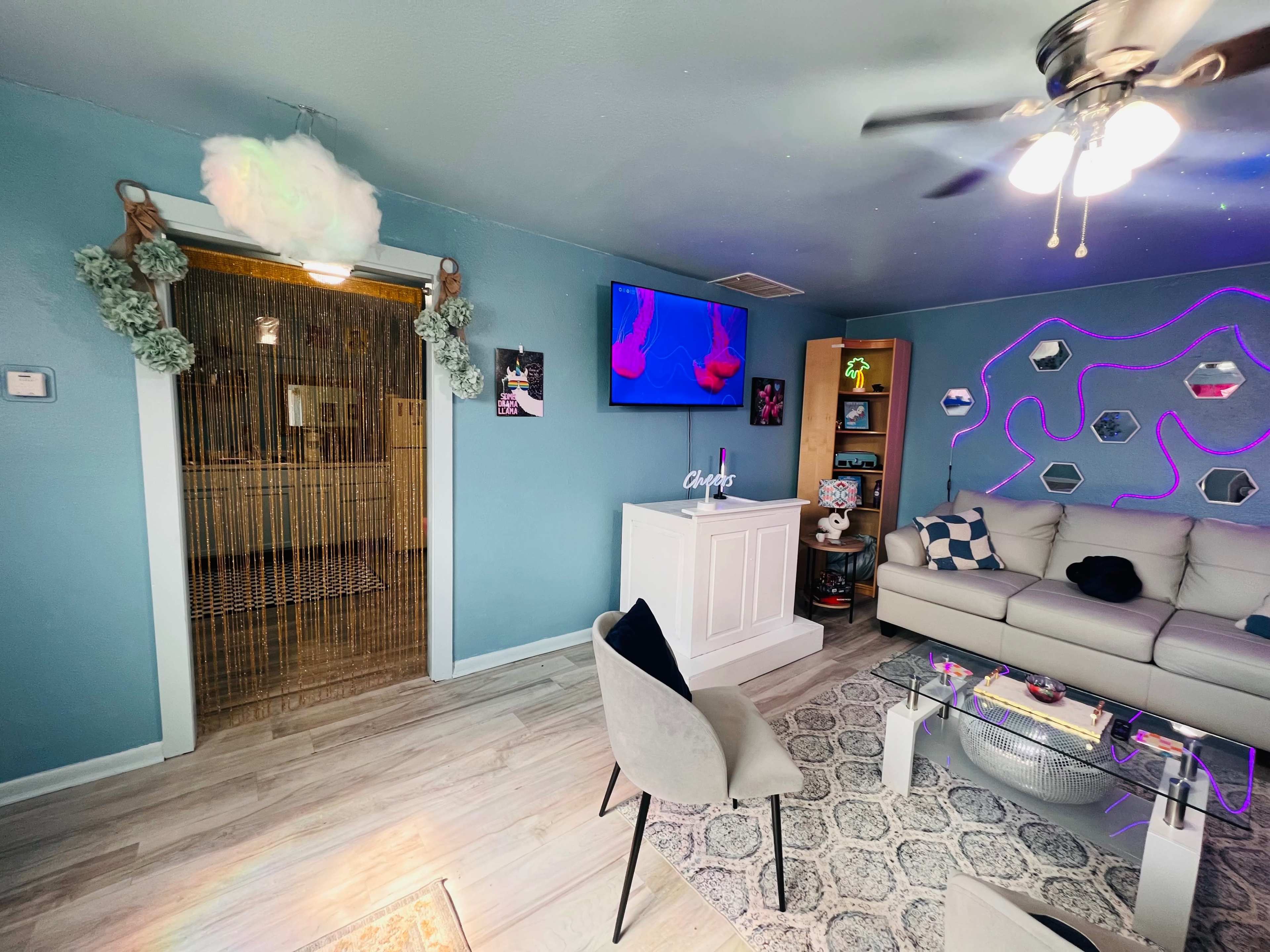 A living room with a light blue wall, a beige sofa, a coffee table, a TV on the wall, and a doorway draped with bamboo curtains leading to another space.