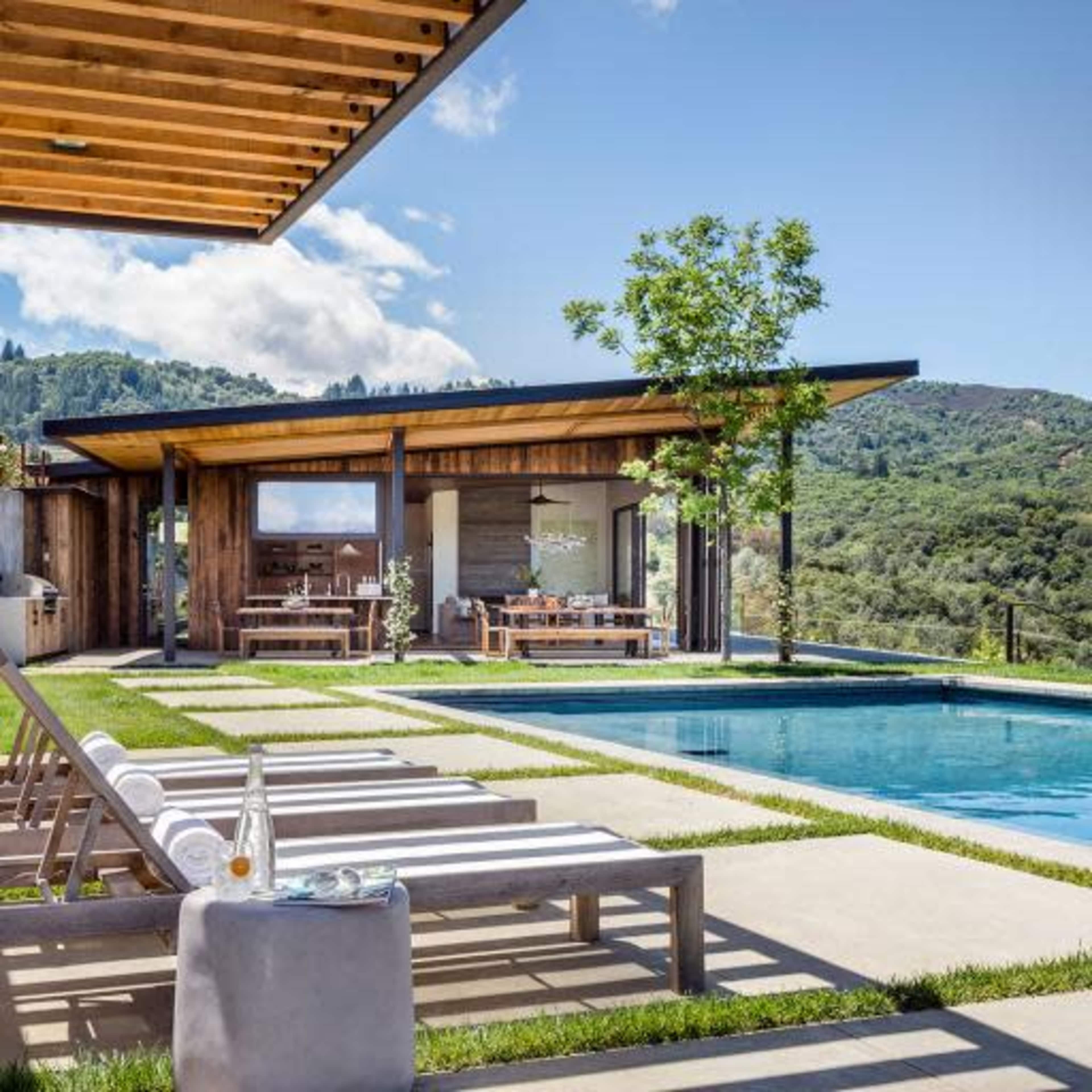 A modern hillside house with a wooden pergola overlooks a swimming pool and landscaped garden.