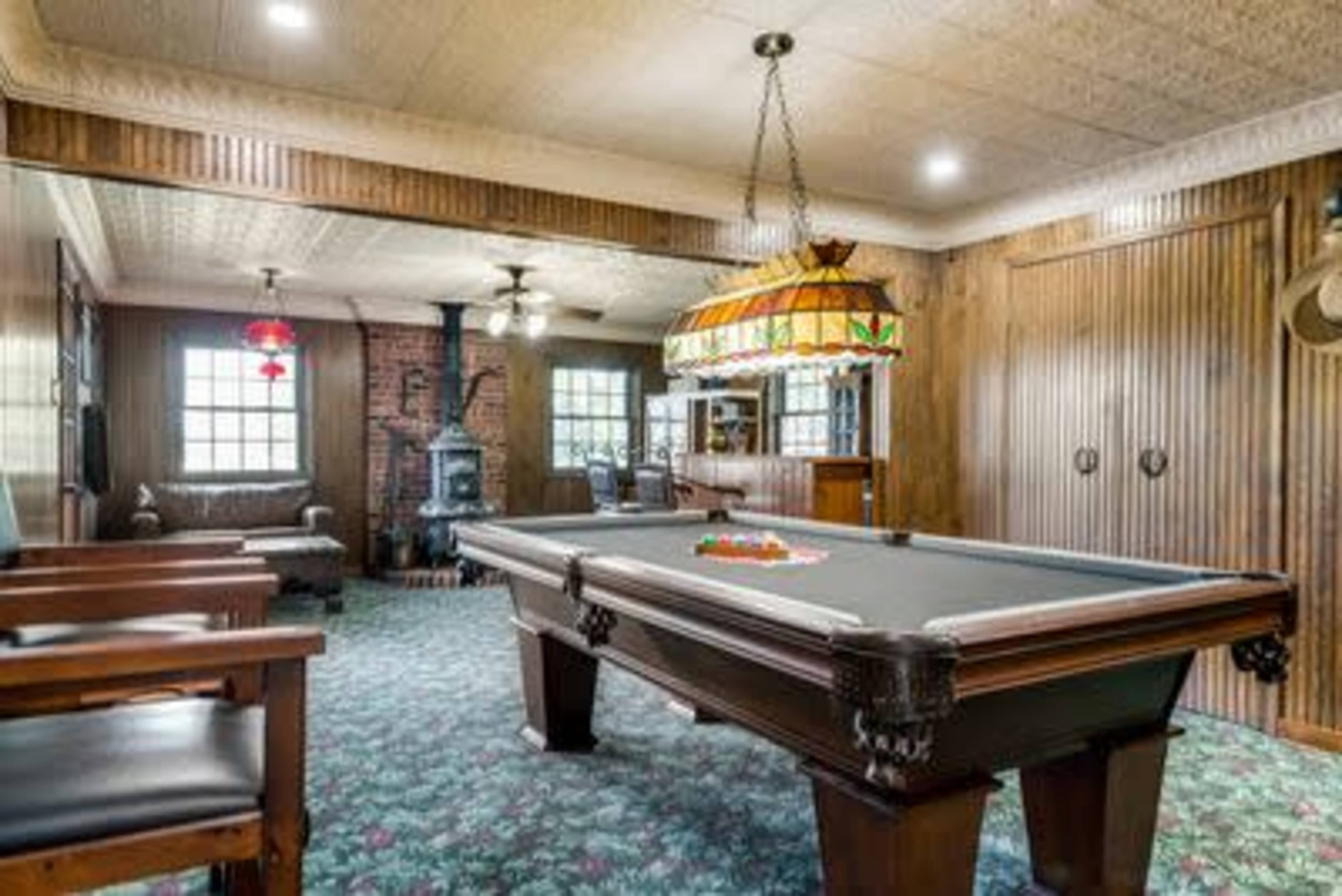 A cozy game room featuring a billiard table, a brick fireplace, wooden paneling, and large windows.