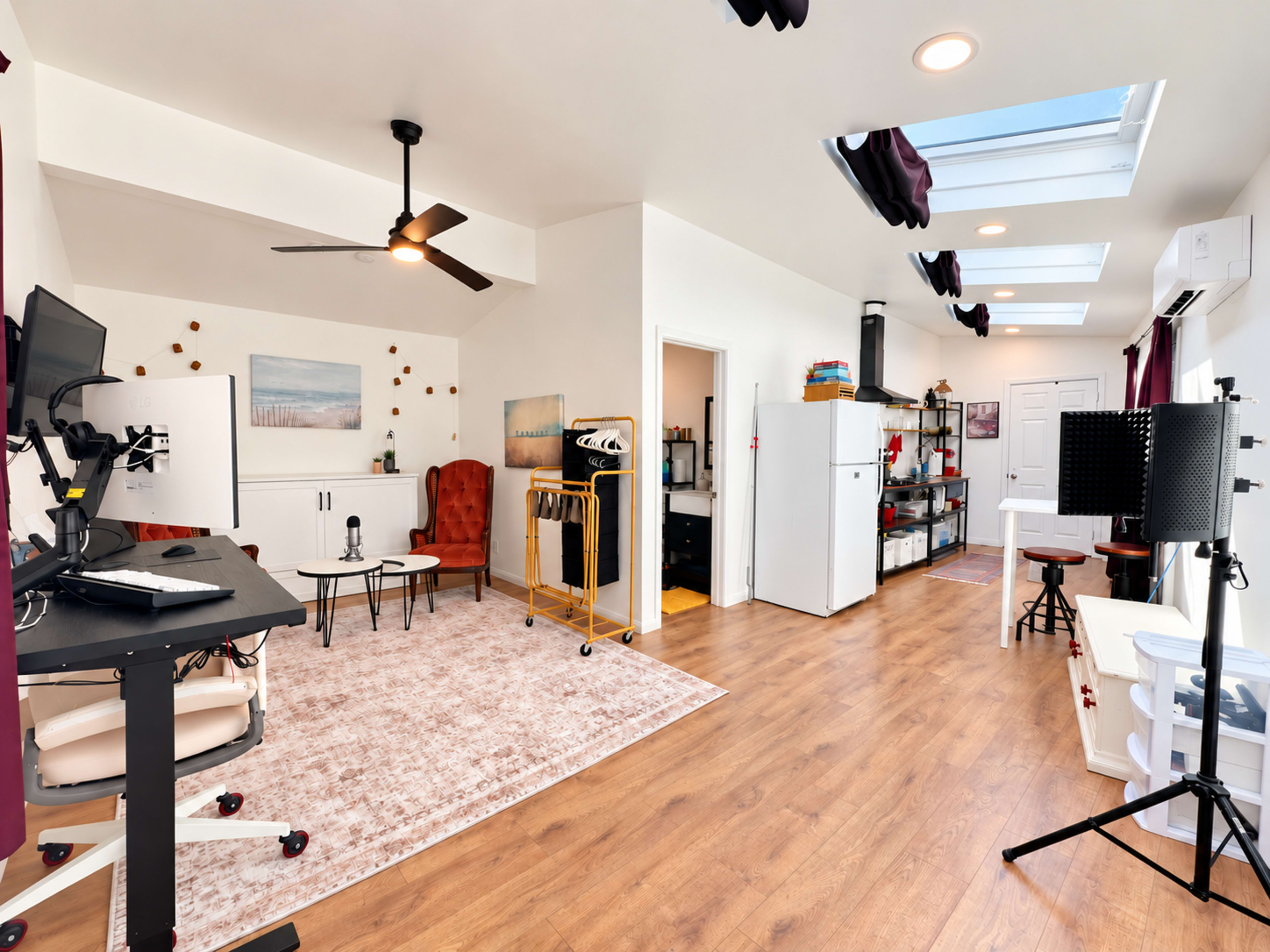 The image shows a spacious, well-lit studio with a mix of furniture, including a desk with monitors, chairs, and a refrigerator, along with skylights allowing natural light to enter.