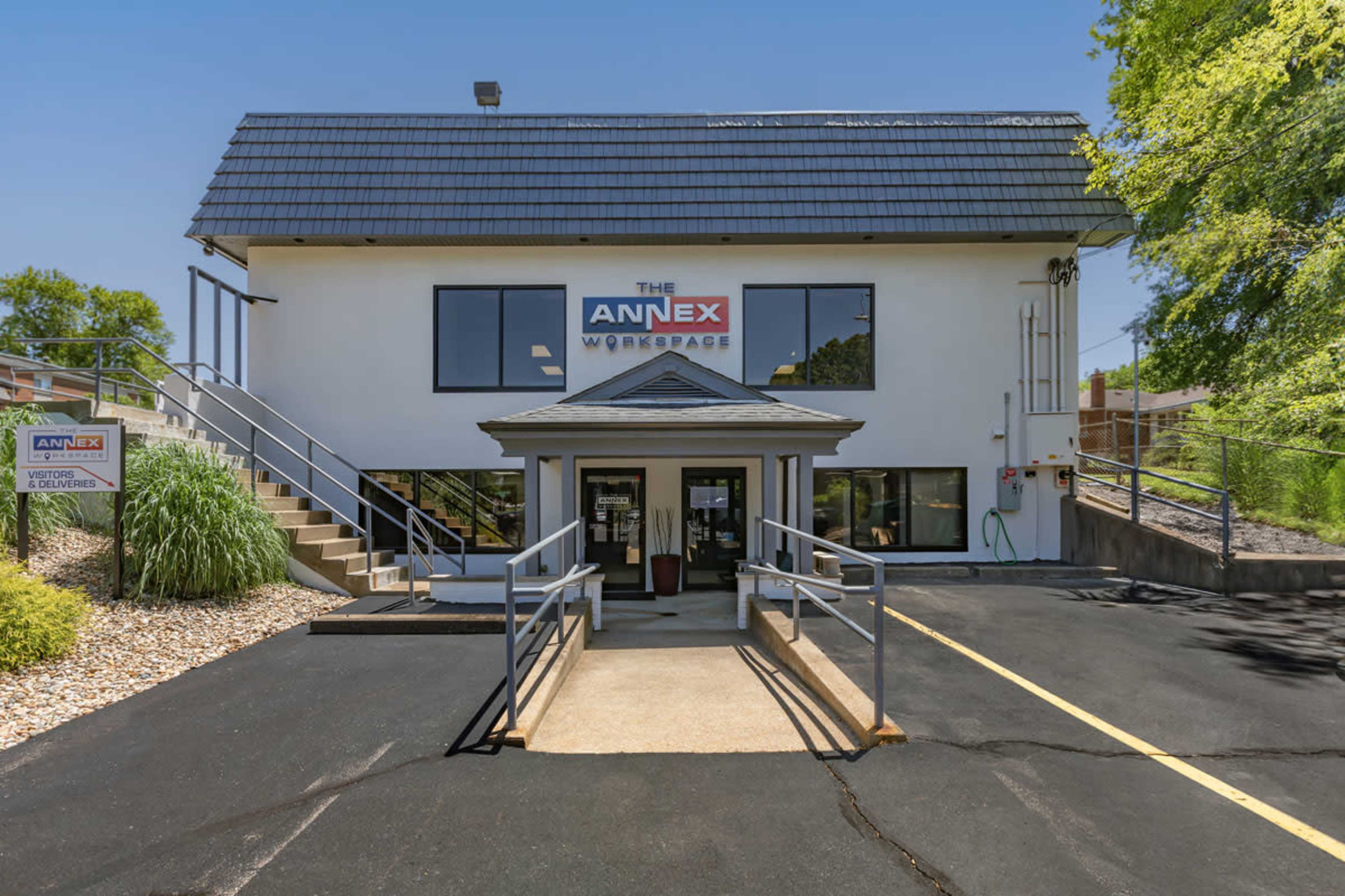 A two-story building with a sloped roof, featuring large windows and a set of steps leading to the entrance, surrounded by landscaped greenery and a paved parking area.
