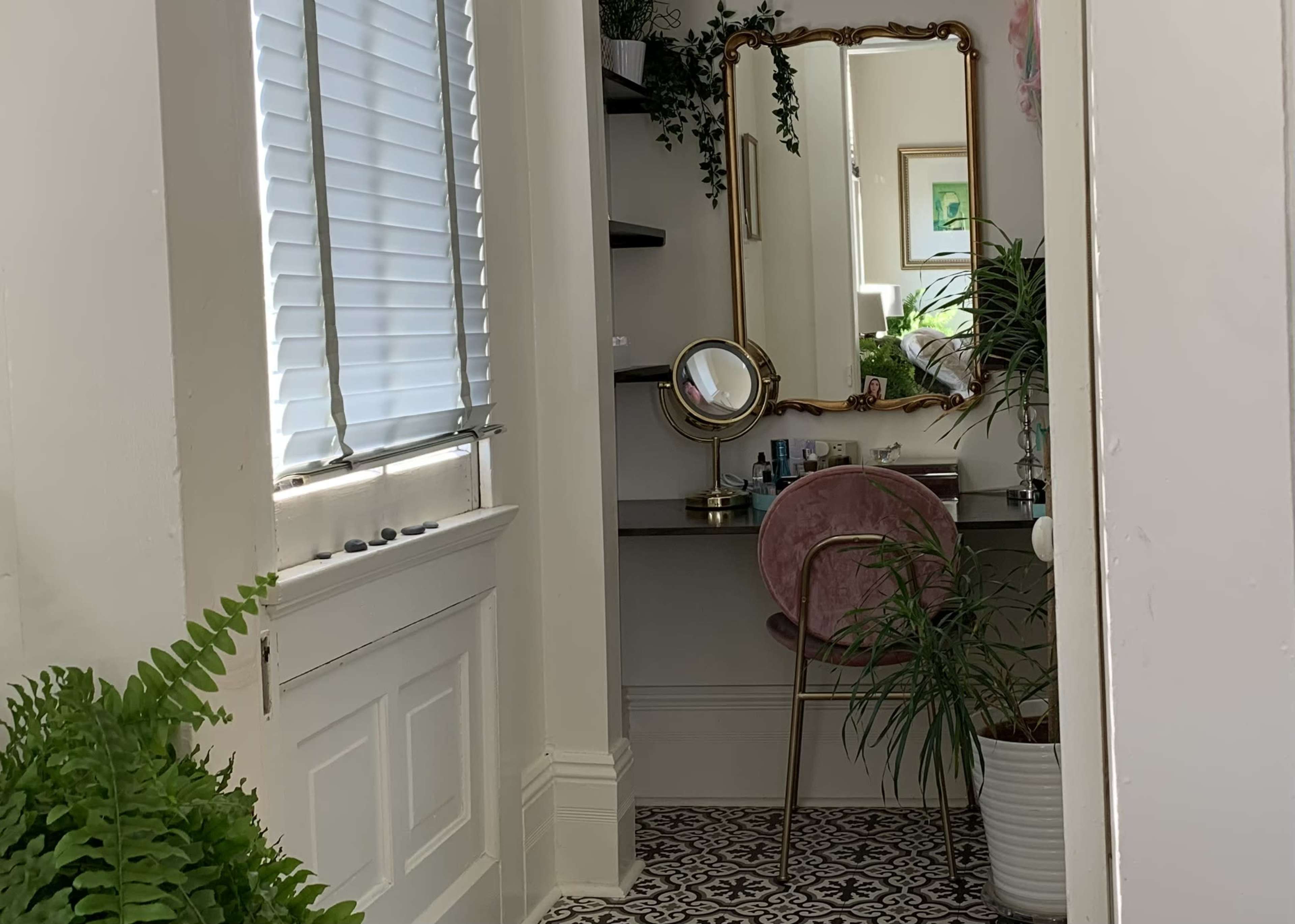 A vanity space with a circular mirror and a pink chair is situated beside a window, adorned with plants and decorative shelves.