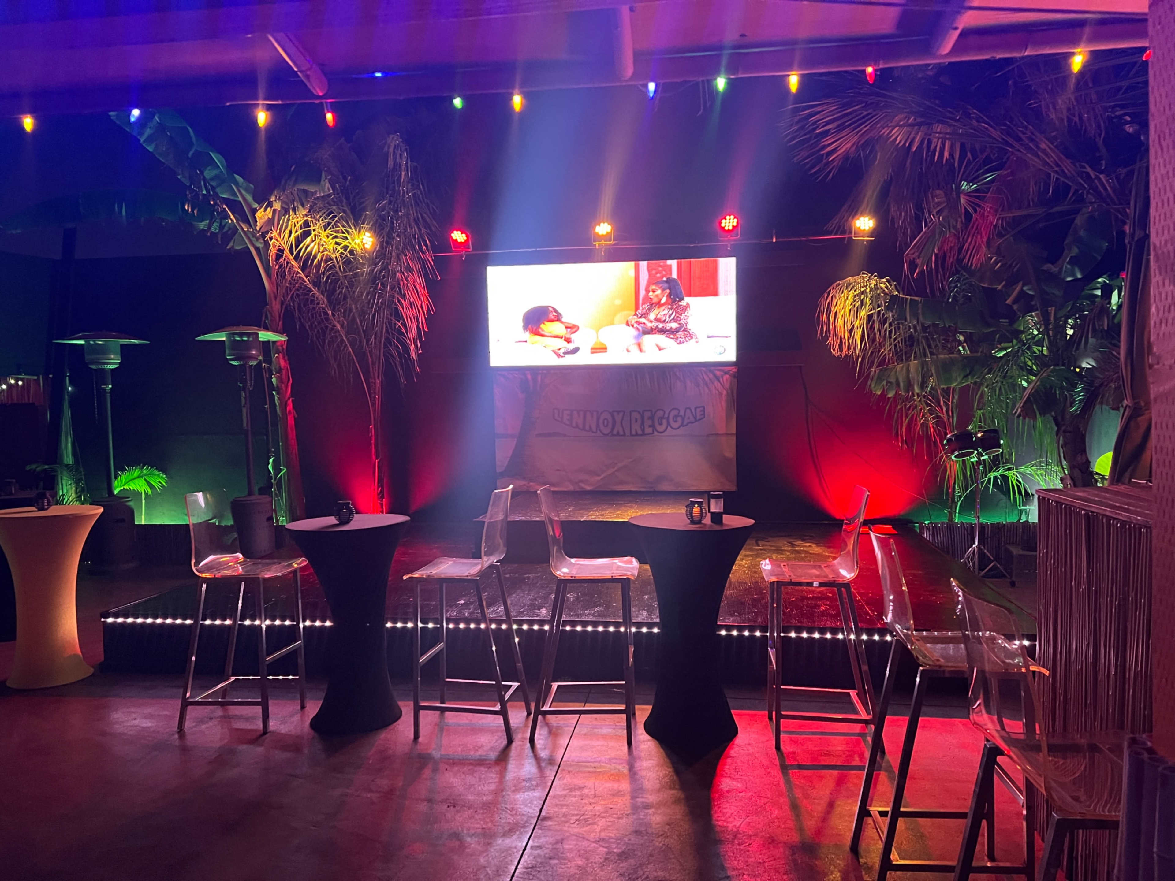 The scene features a dimly lit event space with colorful lights, a large screen displaying content, and several round tables set up for seating.