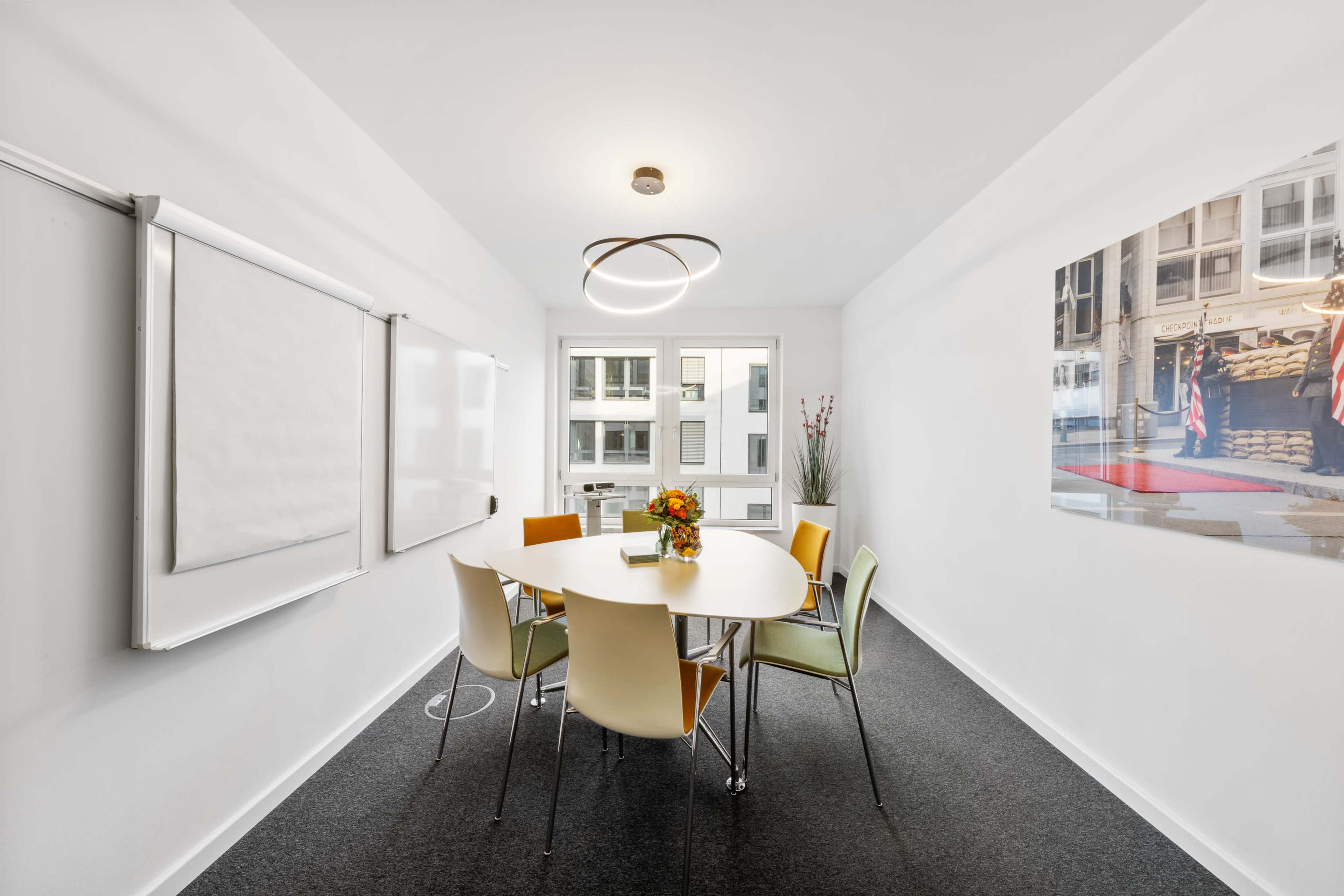 A bright meeting room features a round table surrounded by chairs, with a flower arrangement in the center and a large window illuminating the space.