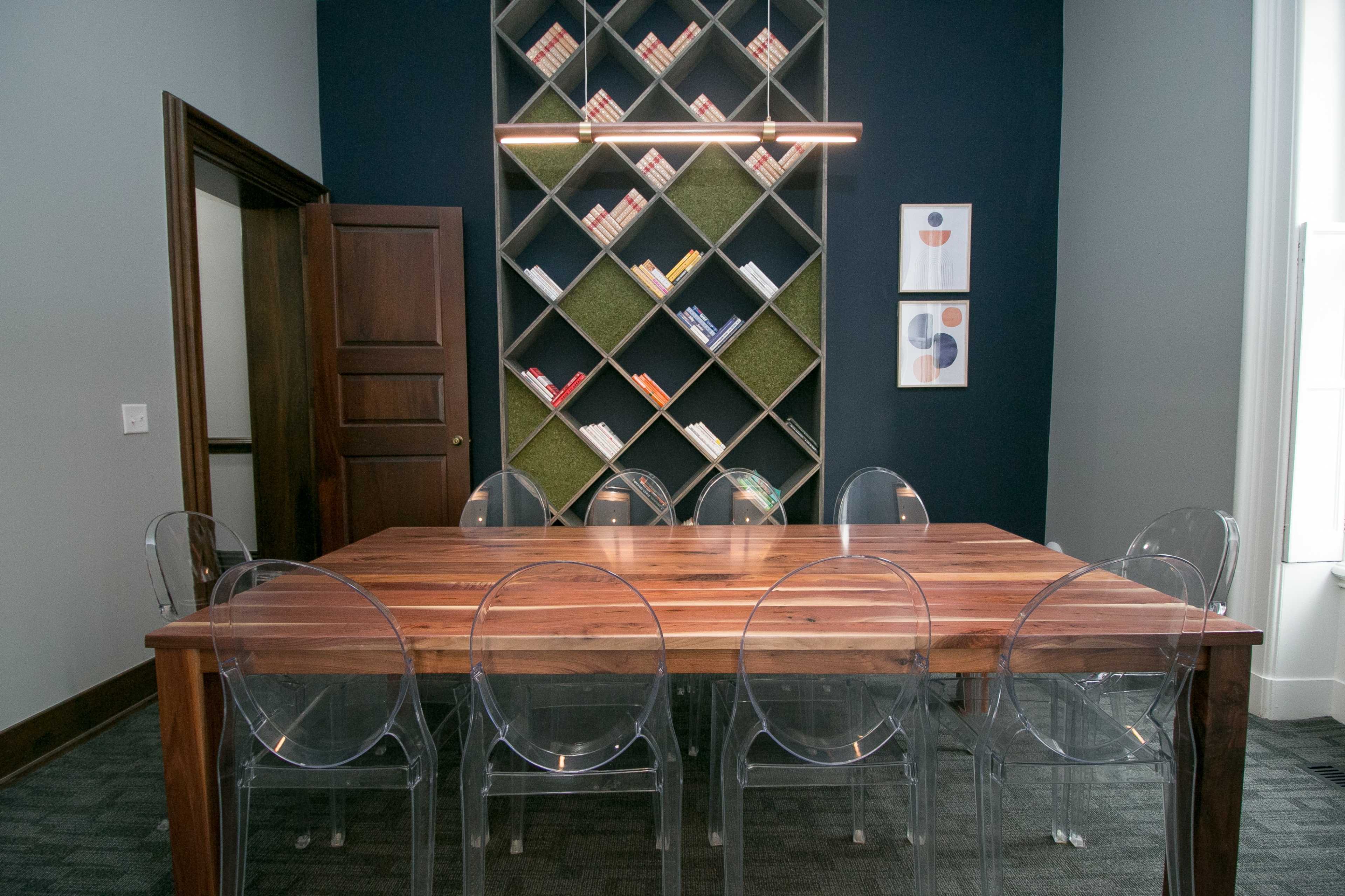 A dining table made of wood is surrounded by clear plastic chairs in a room featuring a bookshelf against a dark wall and two framed artworks.