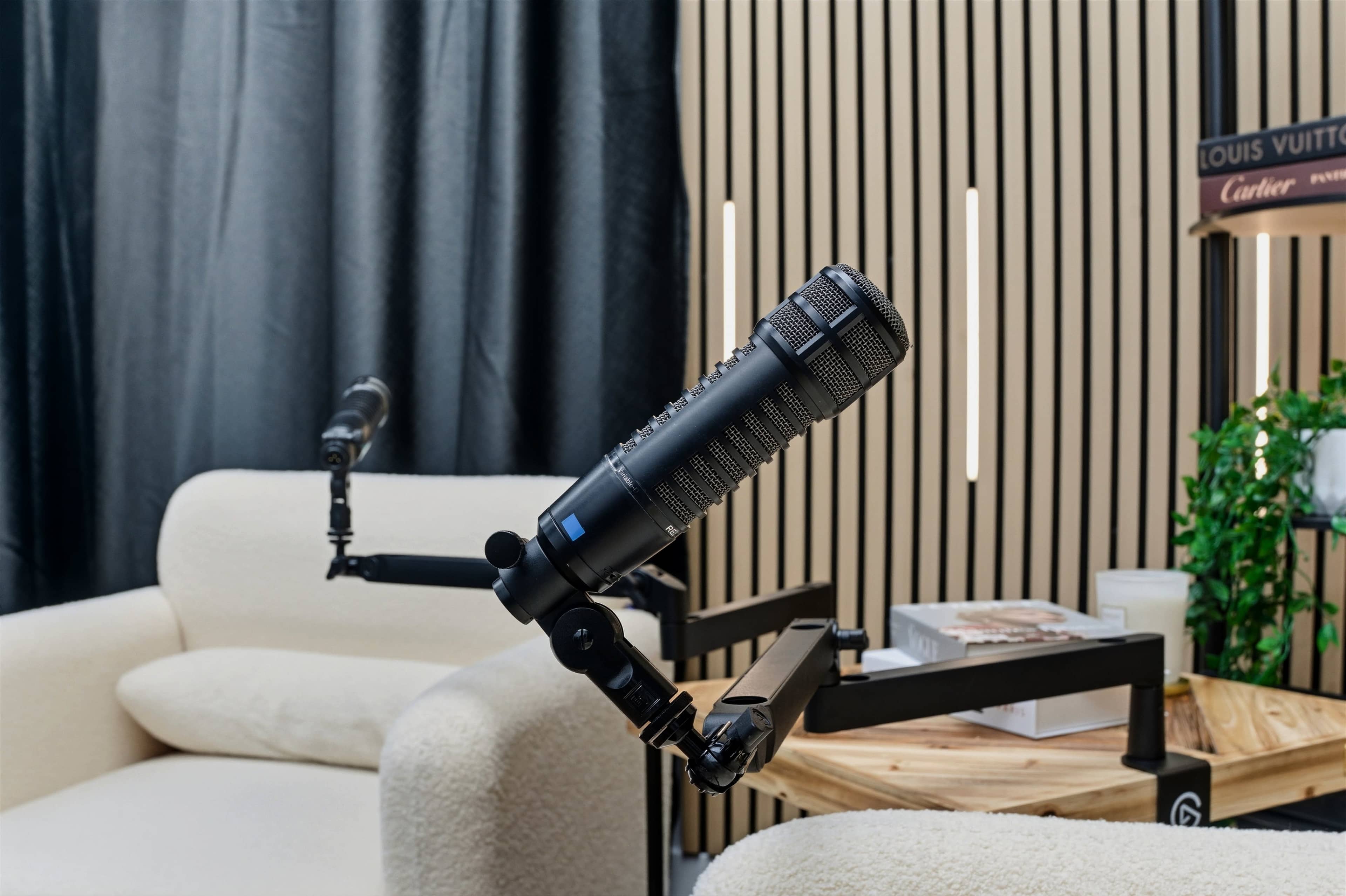 The image shows a black microphone mounted on an adjustable arm, positioned near a light-colored, plush armchair in a cozy interior setting.