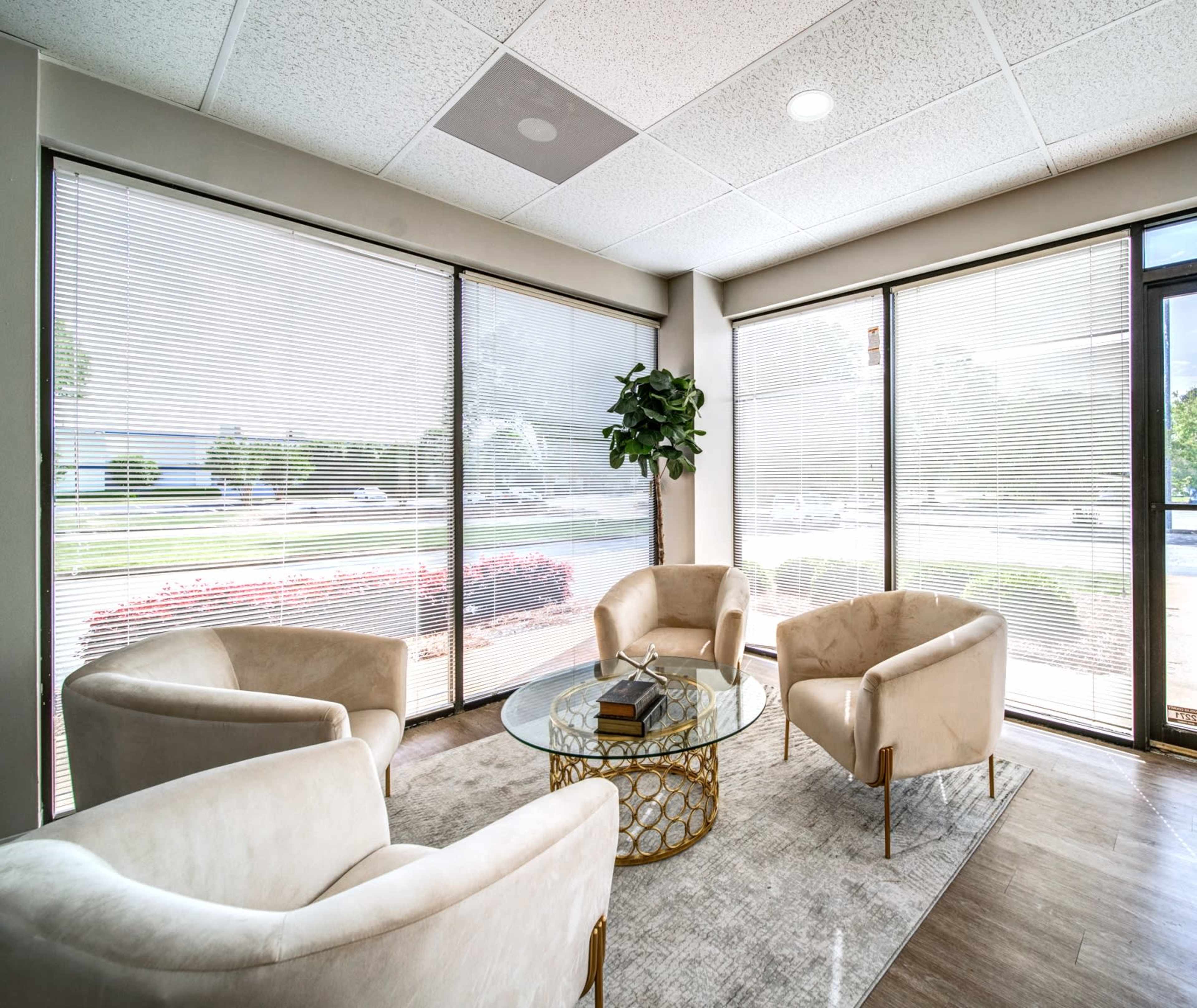 A modern waiting area with three upholstered chairs arranged around a glass coffee table, illuminated by natural light through large windows.