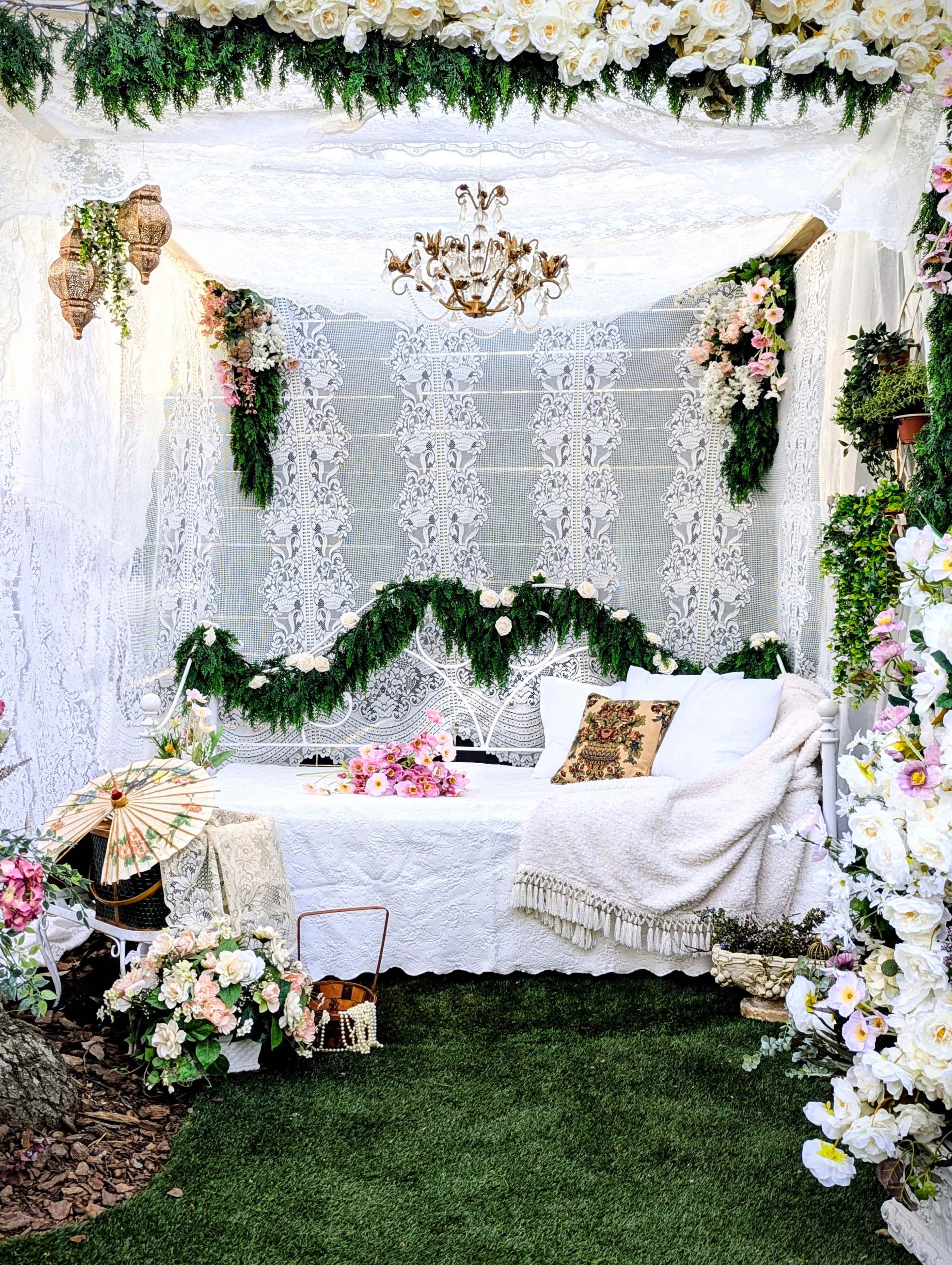 The image shows a decorated outdoor seating area with a lace canopy, floral arrangements, and a white couch surrounded by greenery and decorative elements.