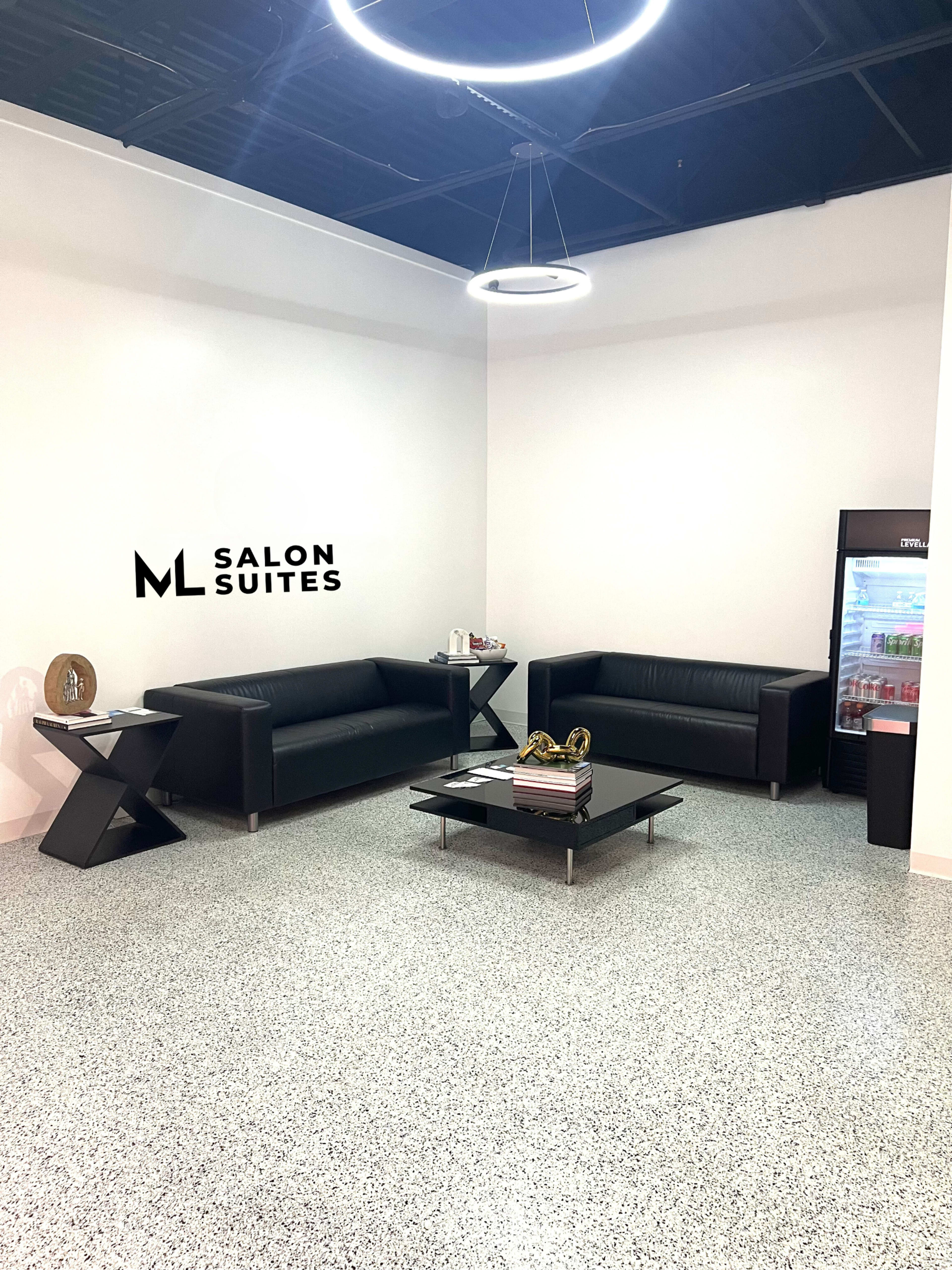 A modern salon suite lobby with two black sofas, a coffee table, and a refrigerator against a white wall featuring the logo "ML SALON SUITES."