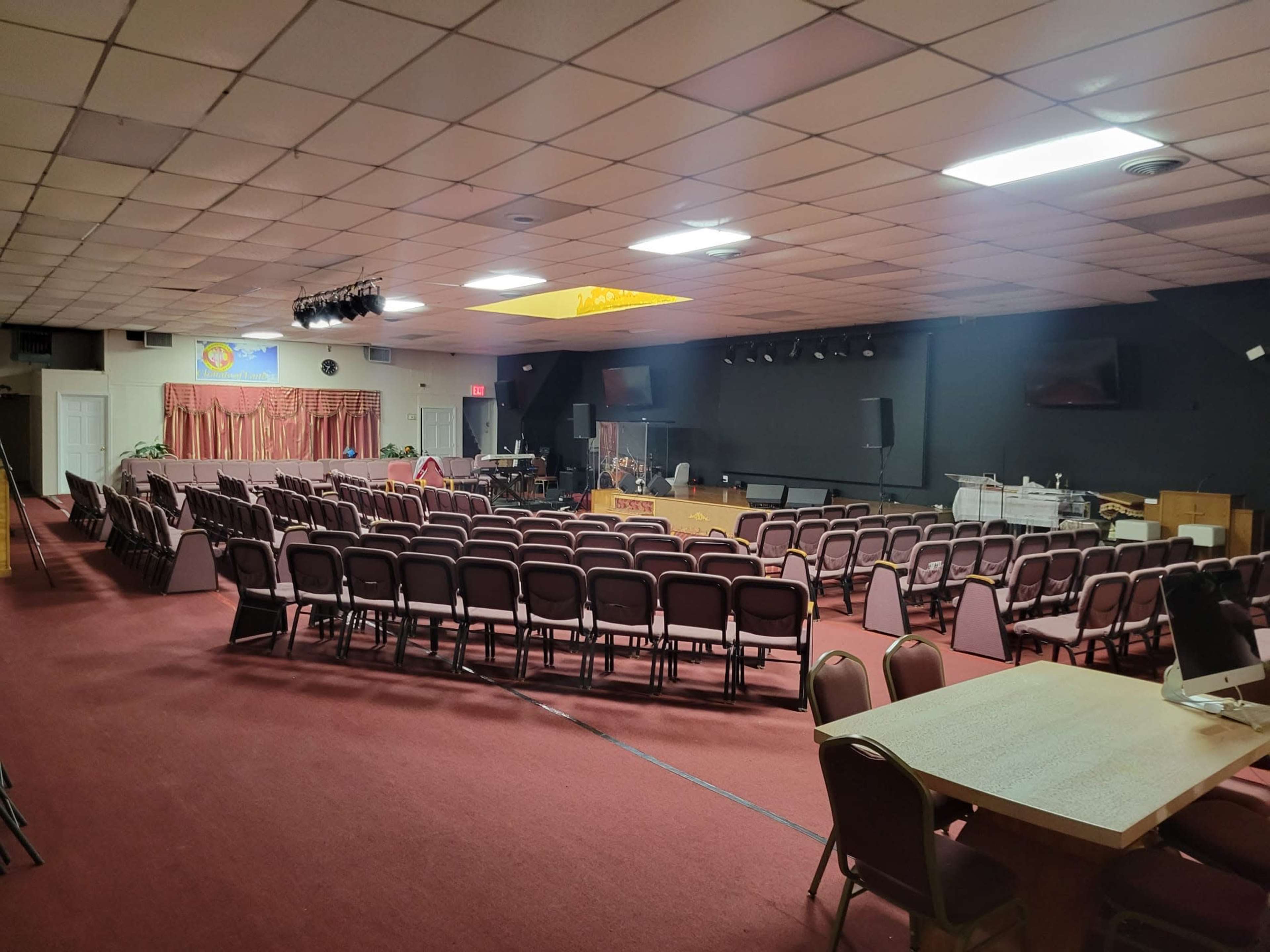 A spacious auditorium set up with rows of chairs facing a stage, with a table and equipment in the corner.