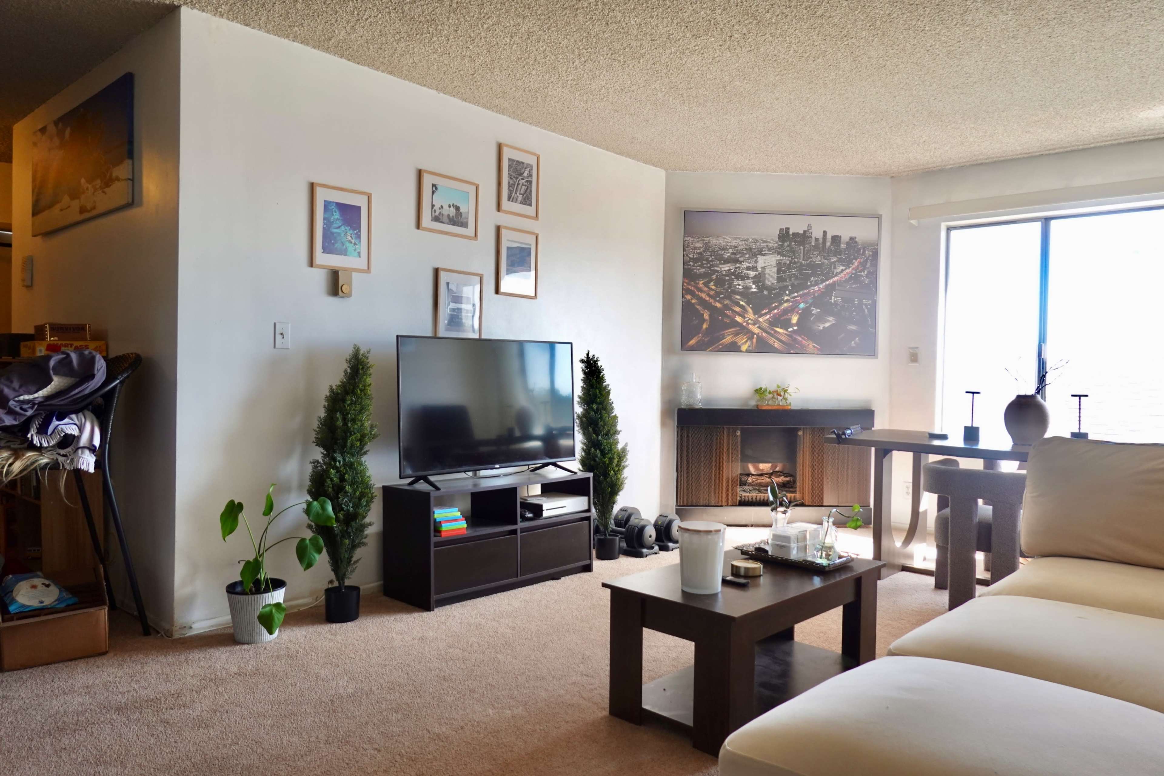 The image shows a cozy living room with a television on a stand, framed pictures on the wall, a small desk, and a sofa facing a large window.