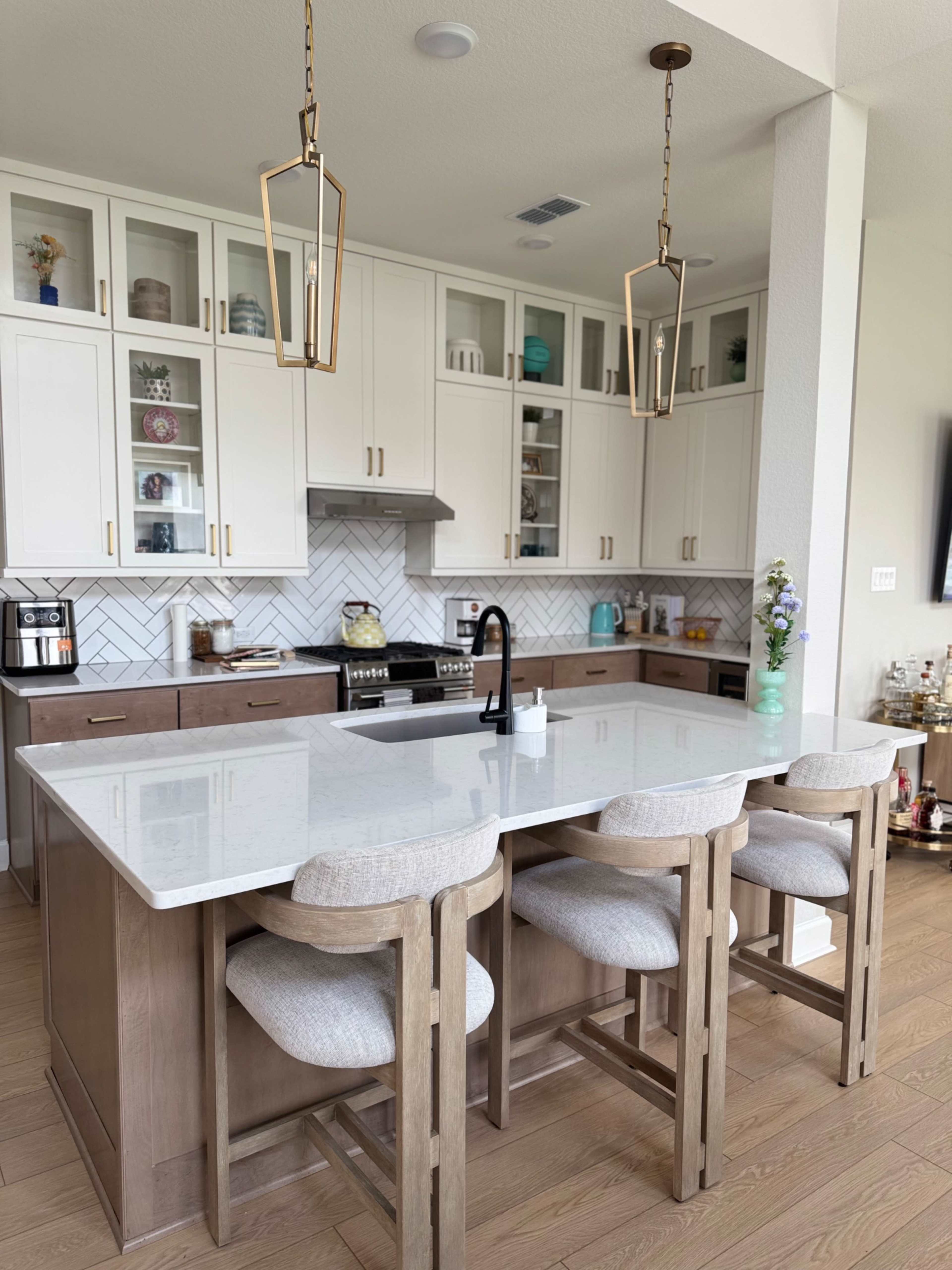 The image shows a modern kitchen featuring white cabinets, a large island with four light-colored stools, and a backsplash made of herringbone-patterned tiles.