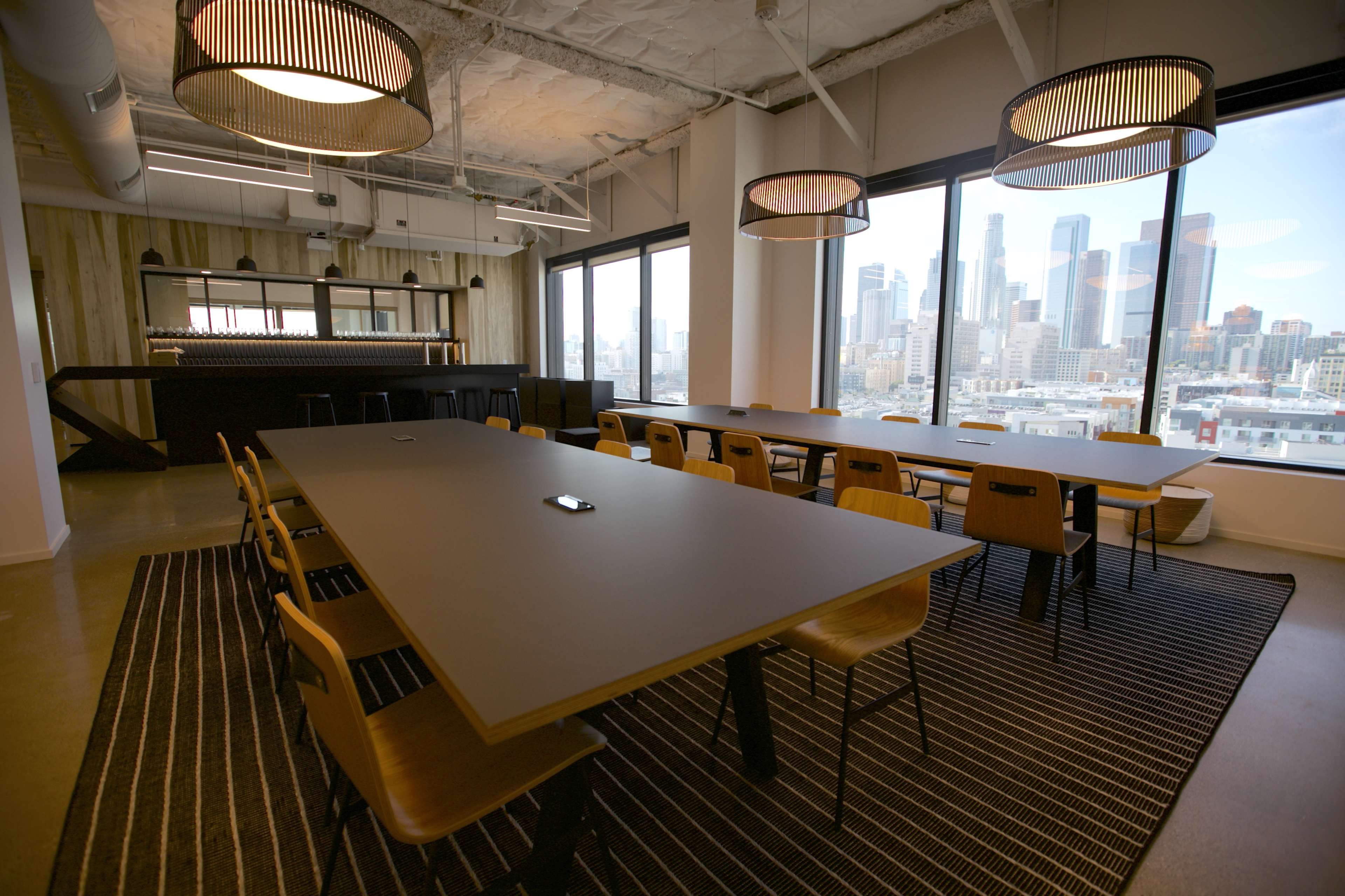 A modern conference room features a long table surrounded by wooden chairs, large windows showcasing a city skyline, and contemporary lighting fixtures.