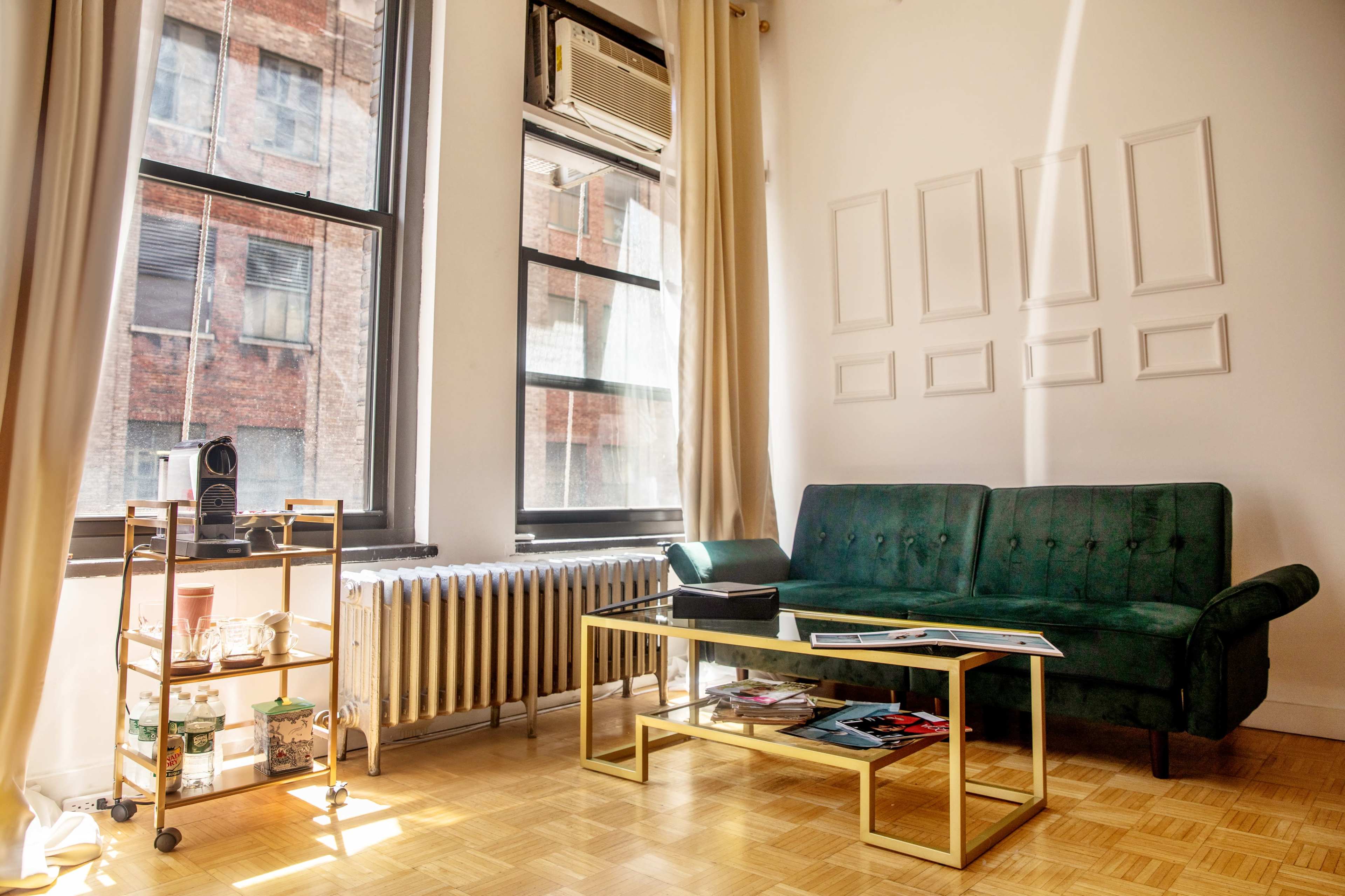 A bright living room with a green sofa, a gold coffee table, and a rolling cart next to a radiator, illuminated by sunlight streaming through large windows.