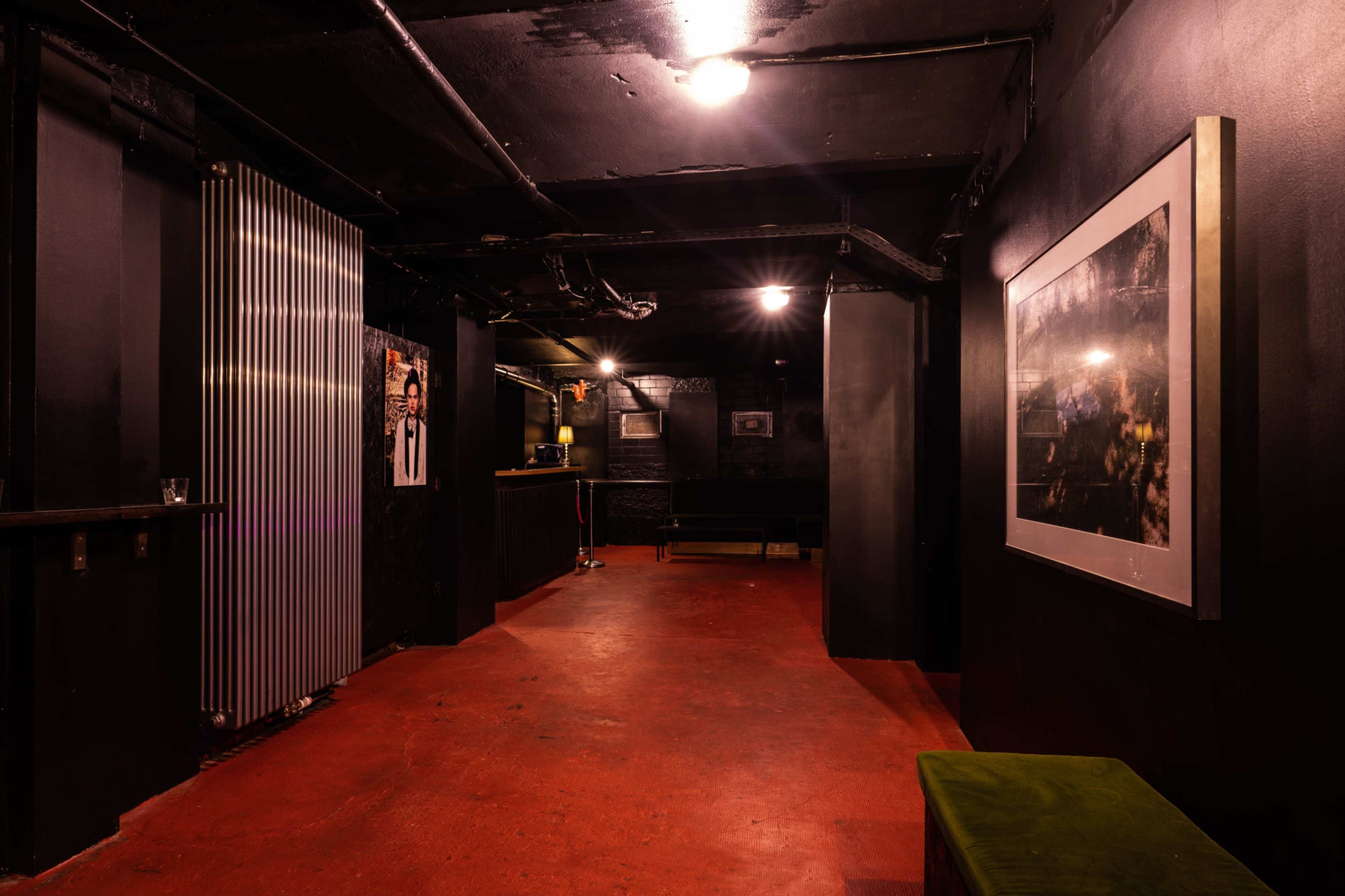 The image shows a dimly lit underground room with a polished red floor, black walls, and a few scattered pieces of furniture, including a bar setup and a large framed photograph.