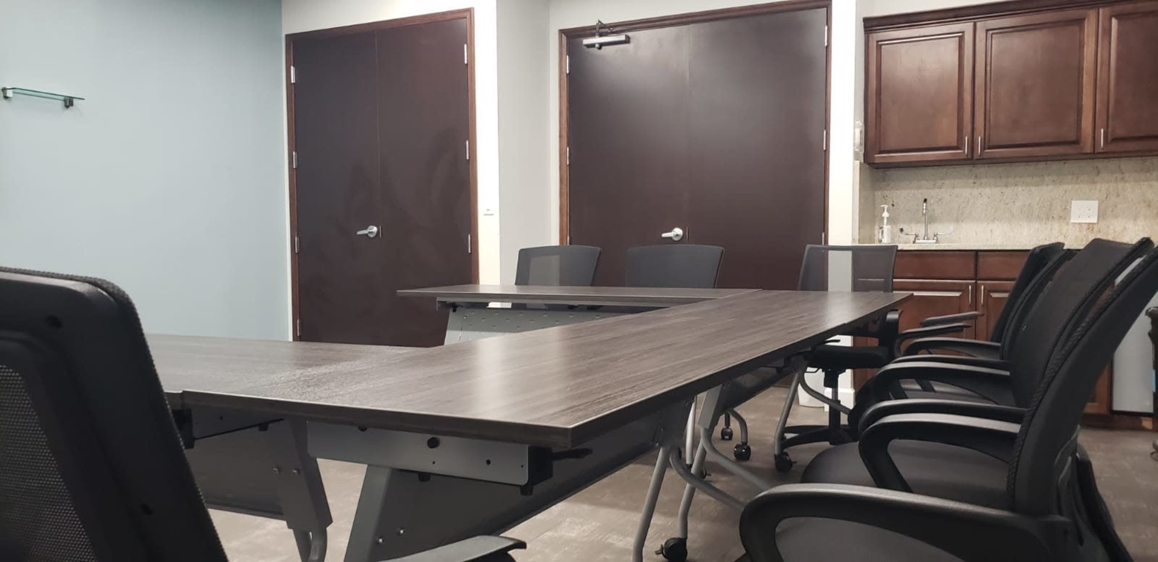 A conference room is equipped with a large table and several black office chairs, featuring a kitchenette in the background.