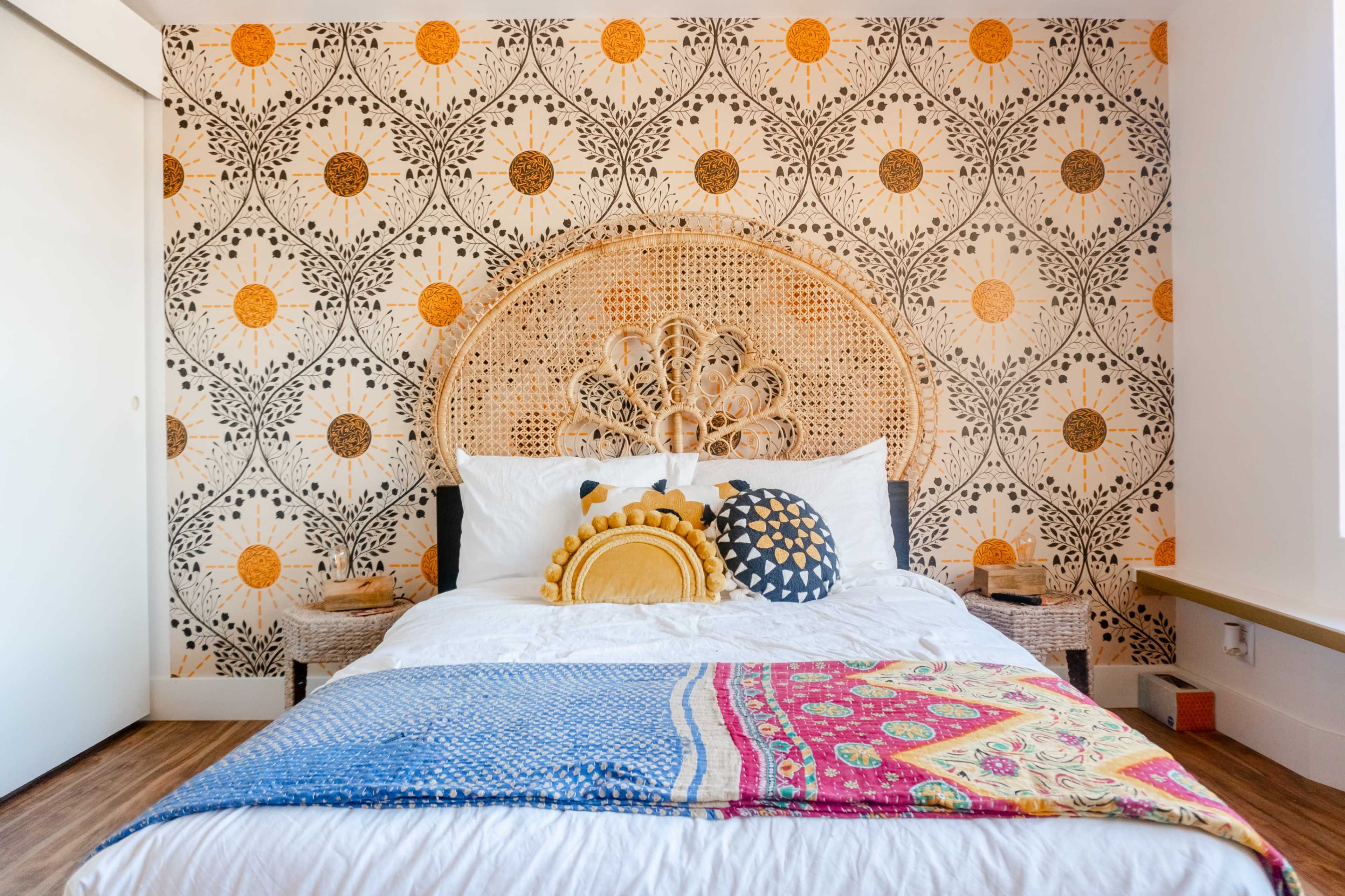 The image shows a bedroom with a large patterned wall, a bed adorned with colorful pillows, and a decorative wicker headboard.