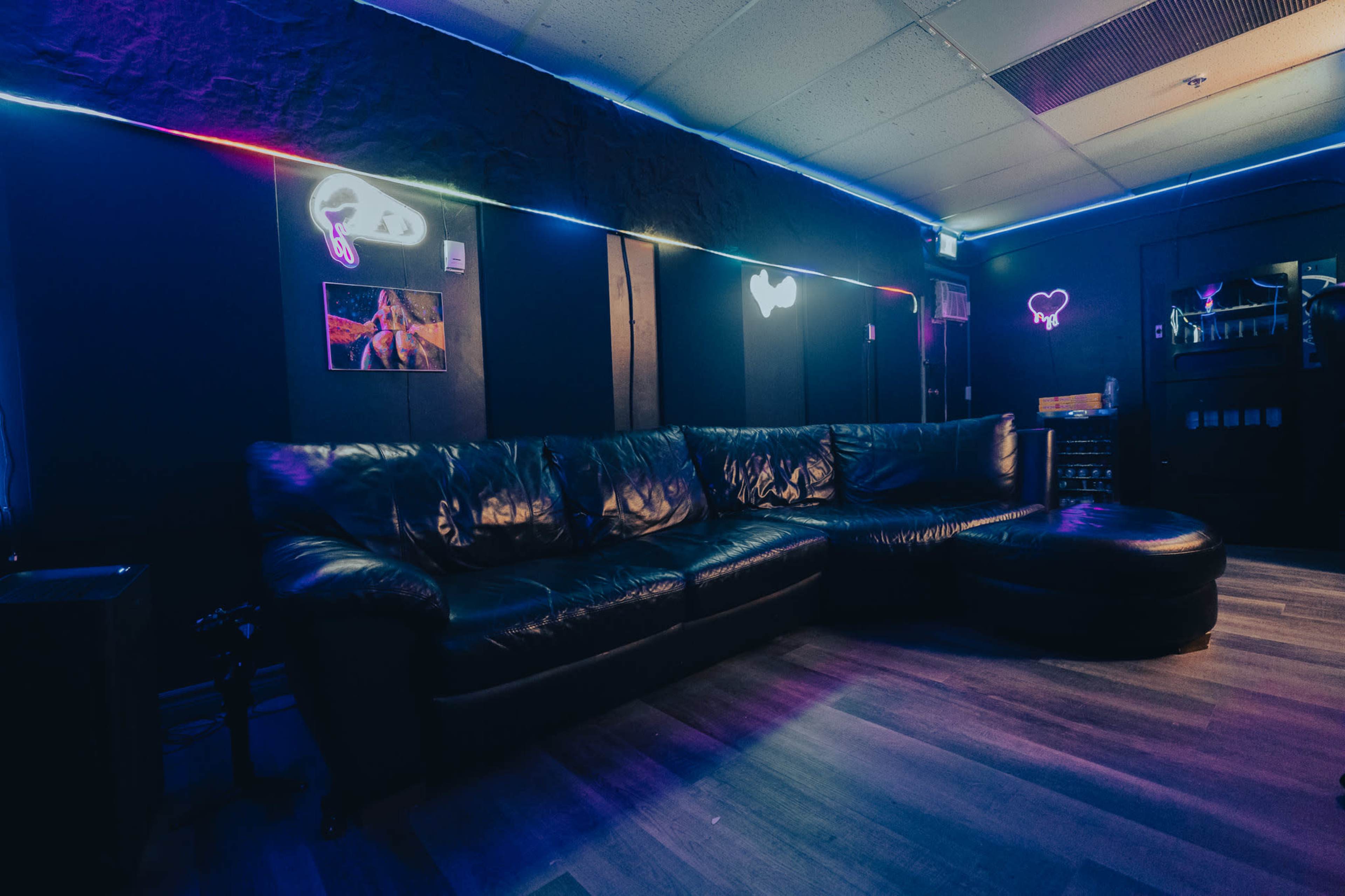 The image shows a dimly lit lounge area featuring a large black leather sofa, a matching ottoman, and neon light accents on the walls.